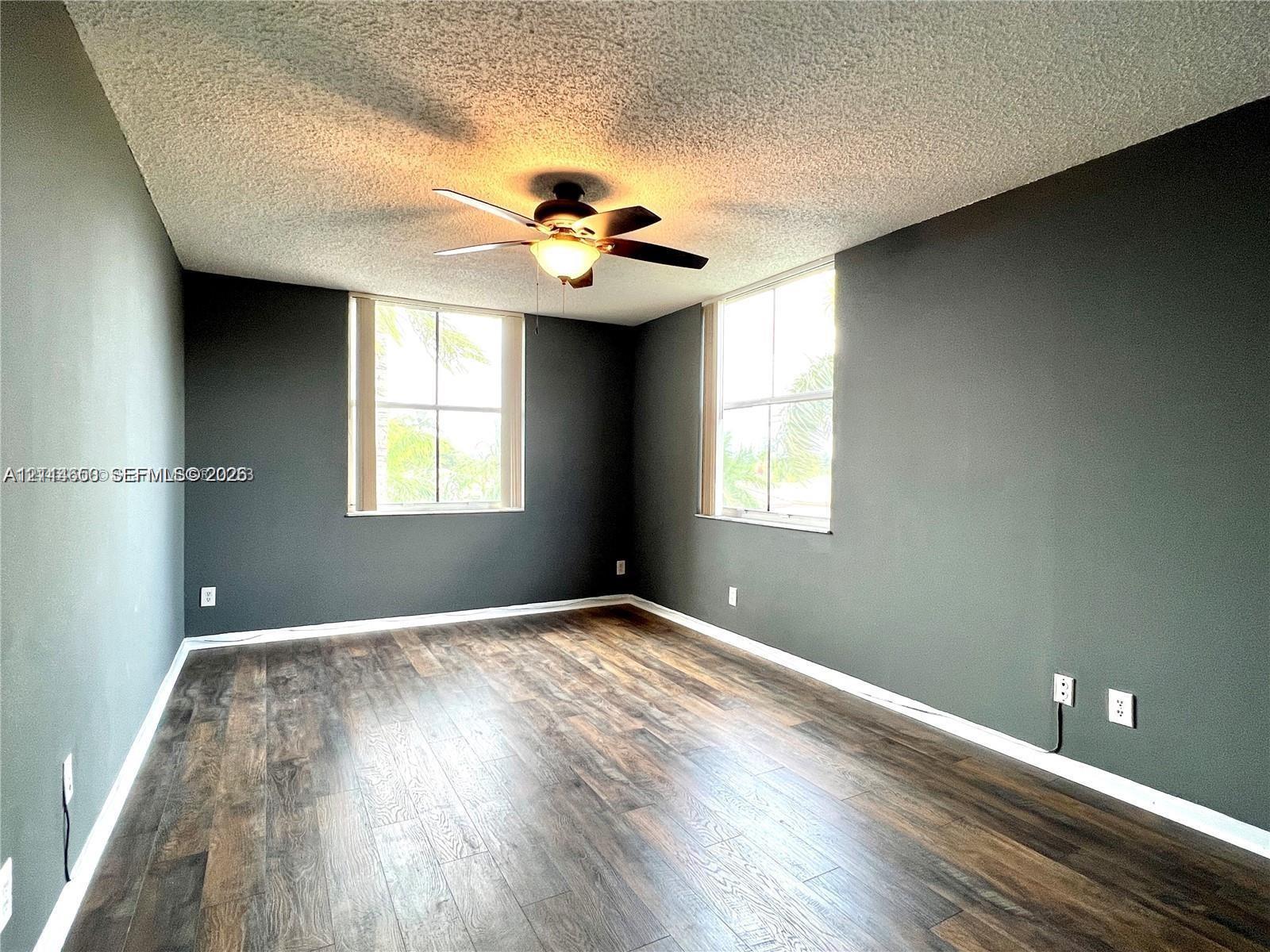 Room featuring dark-toned walls, wood-finish flooring, two large windows, and a central ceiling fan with integrated lighting