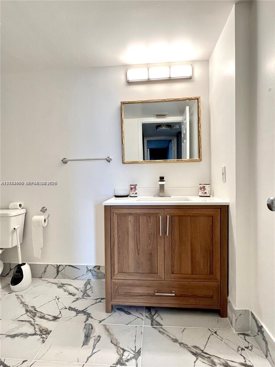 Wood-finish vanity with a rectangular sink, stone-surface countertop, modern wall-mounted mirror, and large-format veined tile flooring