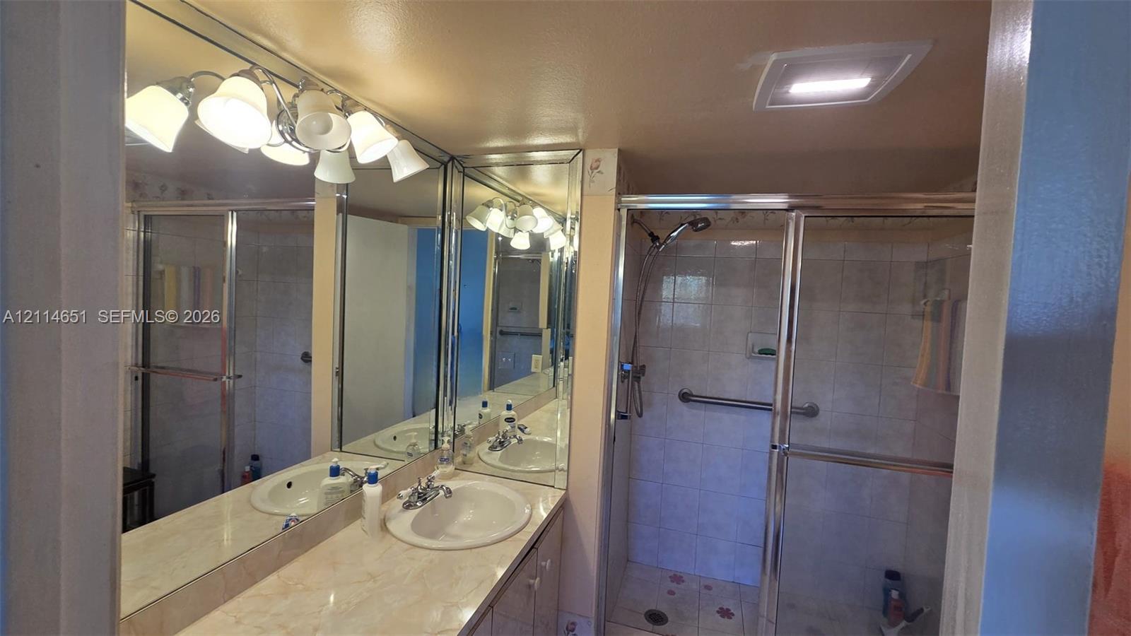 Bathroom vanity featuring dual inset sinks, stone-finish countertops, wall-to-wall mirror, and overhead multi-light fixture, accompanied by a glass-enclosed shower with tiled surround Bathroom vanity featuring dual inset sinks, stone-finish countertops, wall-to-wall mirror, and overhead multi-light fixture, accompanied by a glass-enclosed shower with tiled surround