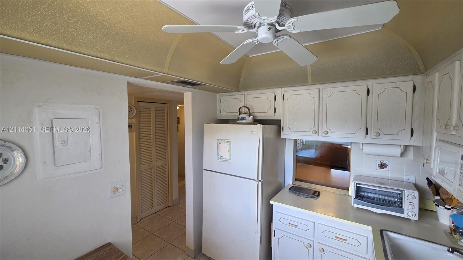 Kitchen featuring white cabinetry with raised-panel doors, neutral solid-surface countertops, tile flooring, a built-in ceiling fan, and curved architectural ceiling detailing Kitchen featuring white cabinetry with raised-panel doors, neutral solid-surface countertops, tile flooring, a built-in ceiling fan, and curved architectural ceiling detailing