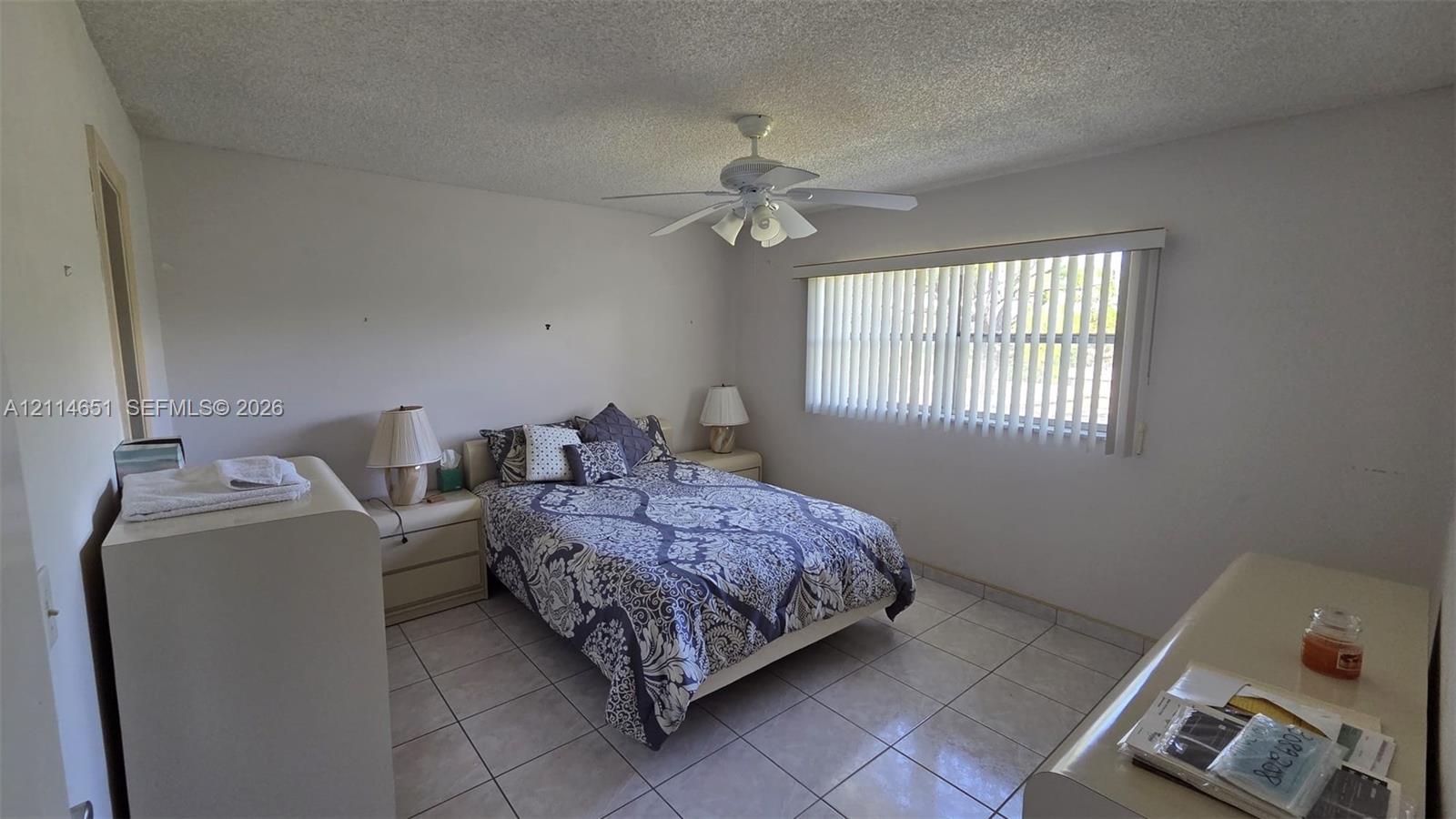 Room featuring light-colored tile flooring, a multi-blade ceiling fan, and vertical window blinds Room featuring light-colored tile flooring, a multi-blade ceiling fan, and vertical window blinds