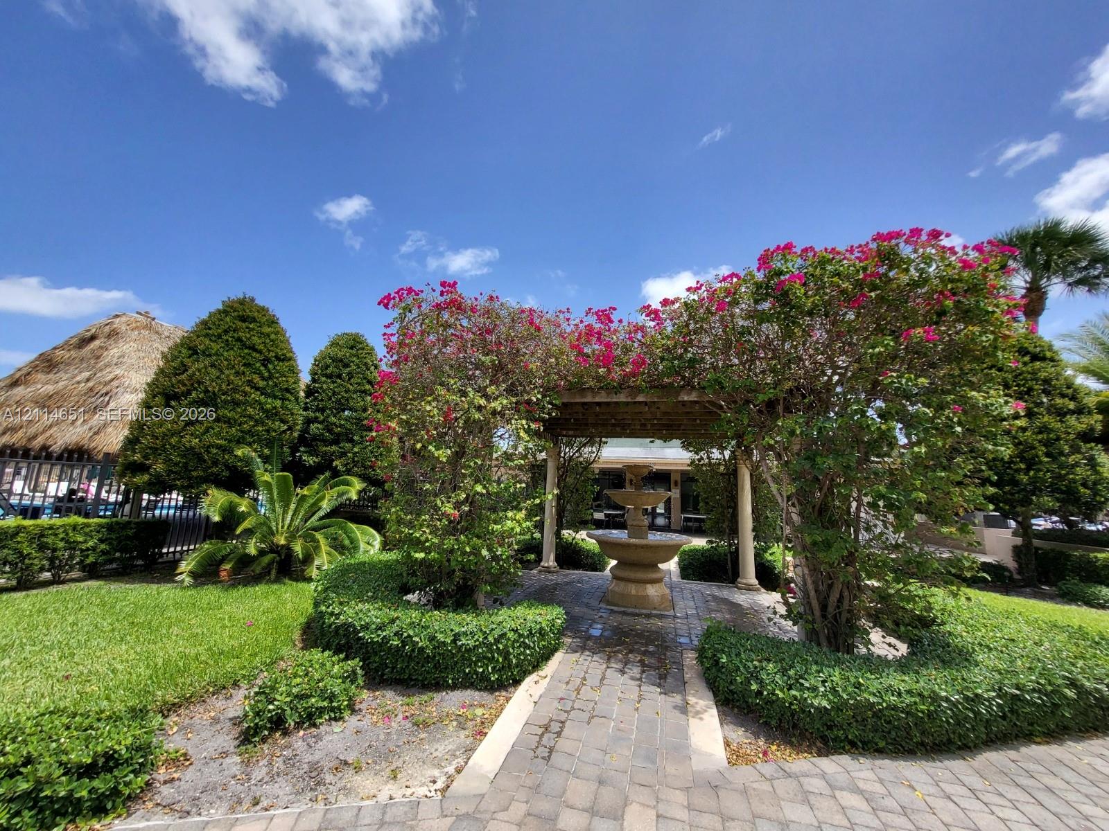 Outdoor garden featuring a tiered stone fountain, stone paver pathway, wooden pergola, and sculpted hedge landscaping