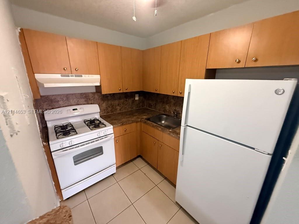 Kitchen featuring wood-finish cabinetry, stone-texture laminate countertops, a stainless steel sink, a white gas range, and ceramic tile flooring