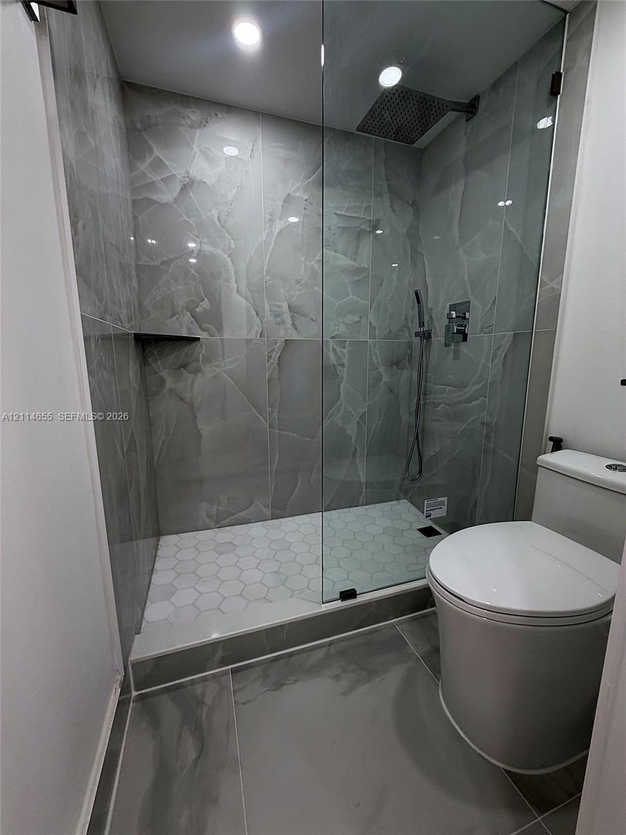 Walk-in shower featuring large-format stone-look wall tiling, white hexagonal floor tiling, a ceiling-mounted rainfall showerhead, a wall-mounted handheld sprayer, and a recessed corner shelf