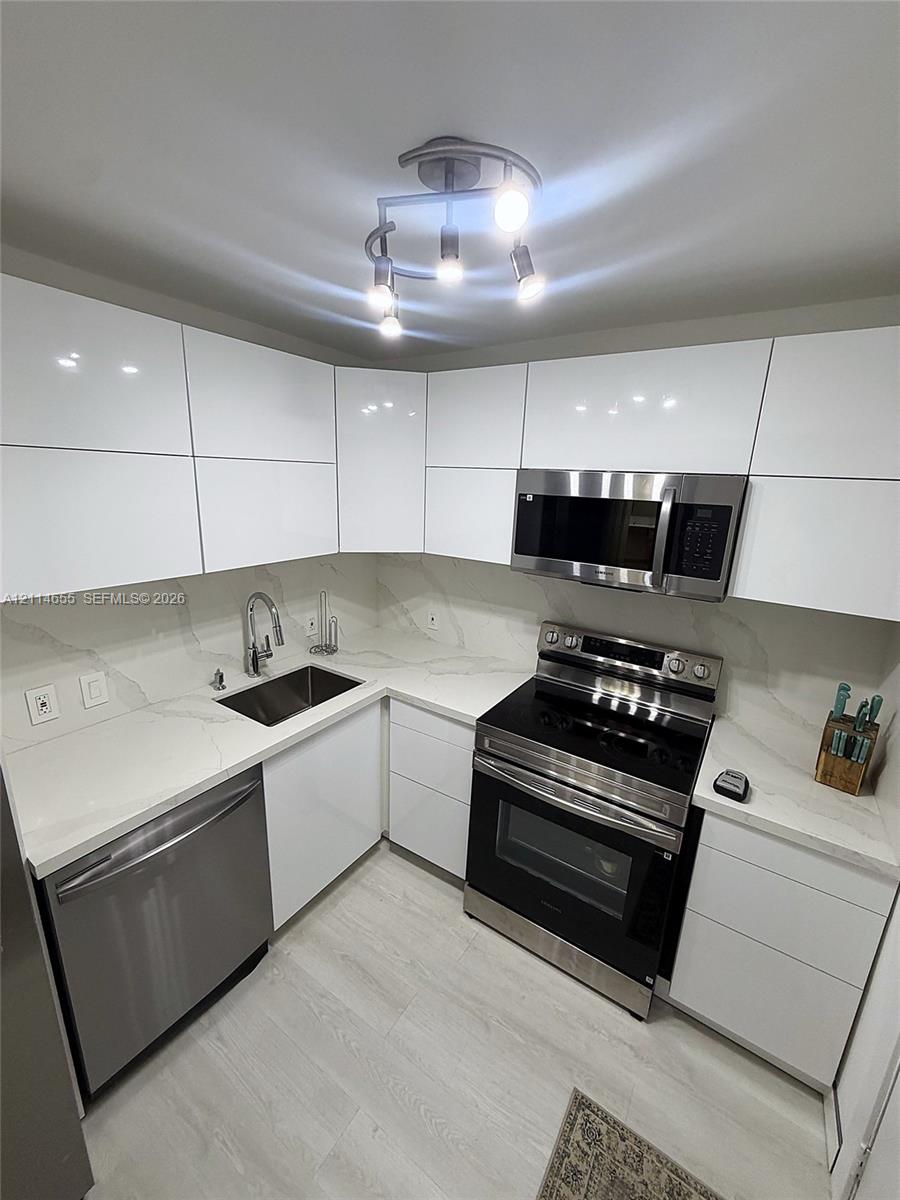 Modern kitchen featuring high-gloss white flat-panel cabinetry, light-toned stone-finish countertops, stainless steel appliances, and a contemporary multi-bulb ceiling fixture