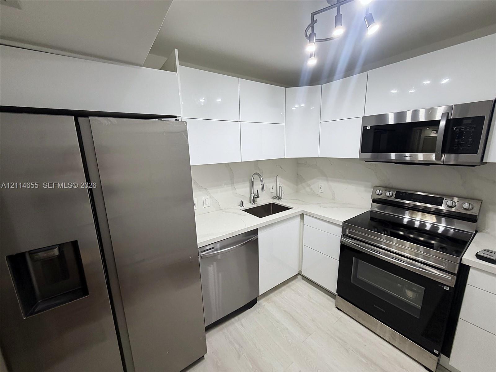 Kitchen featuring high-gloss white cabinetry, white stone-finish countertops, full-height stone-finish backsplash, stainless steel appliances, and wood-finish flooring