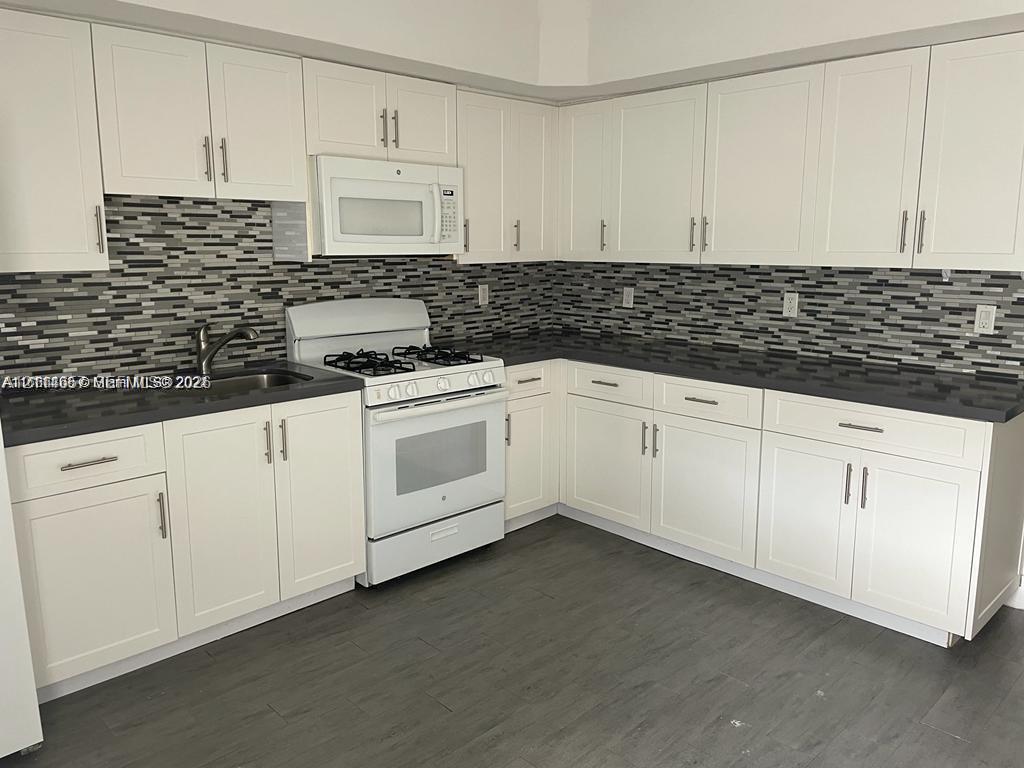 Kitchen featuring white shaker-style cabinetry, dark stone-finish countertops, linear tile backsplash, wood-finish flooring, and white built-in appliances