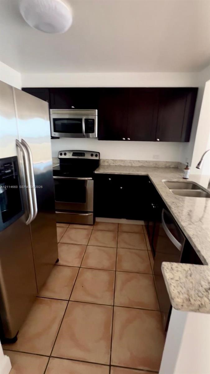 Kitchen featuring dark-finish cabinetry, stone-style countertops, ceramic tile flooring, and stainless steel appliances including a built-in microwave, range, and double-door refrigerator Kitchen featuring dark-finish cabinetry, stone-style countertops, ceramic tile flooring, and stainless steel appliances including a built-in microwave, range, and double-door refrigerator