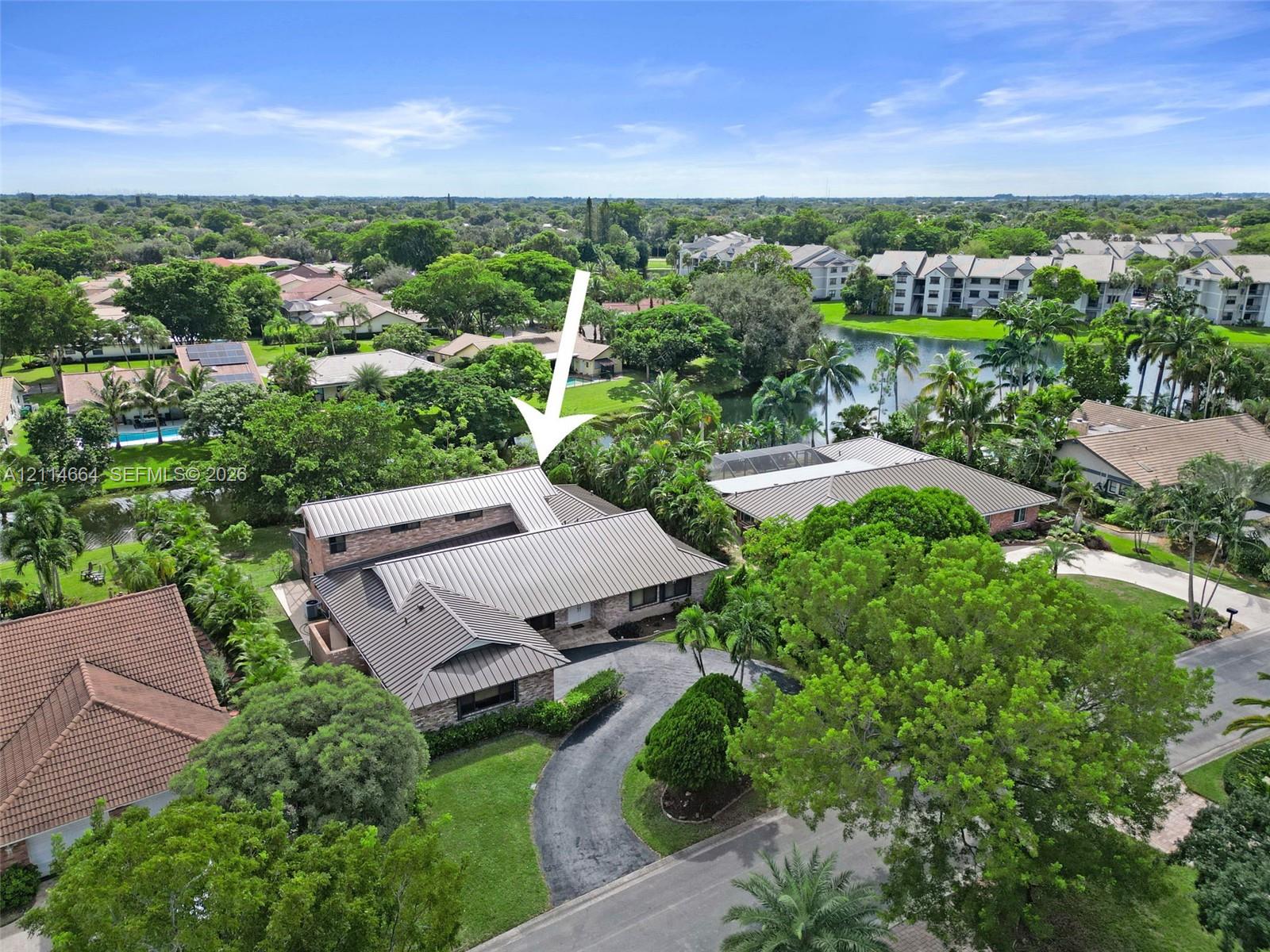 Aerial view featuring a multi-pitched metal roof, brick exterior cladding, curved driveway, and lush perimeter landscaping situated along a waterway