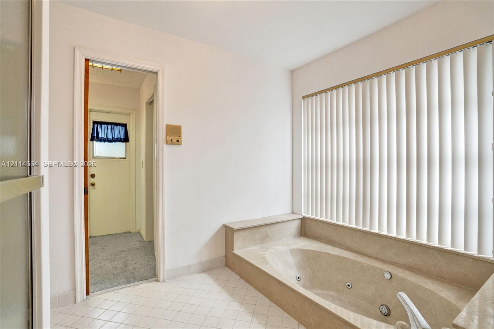 Built-in jetted tub with surround, large window with vertical blinds, and tile flooring