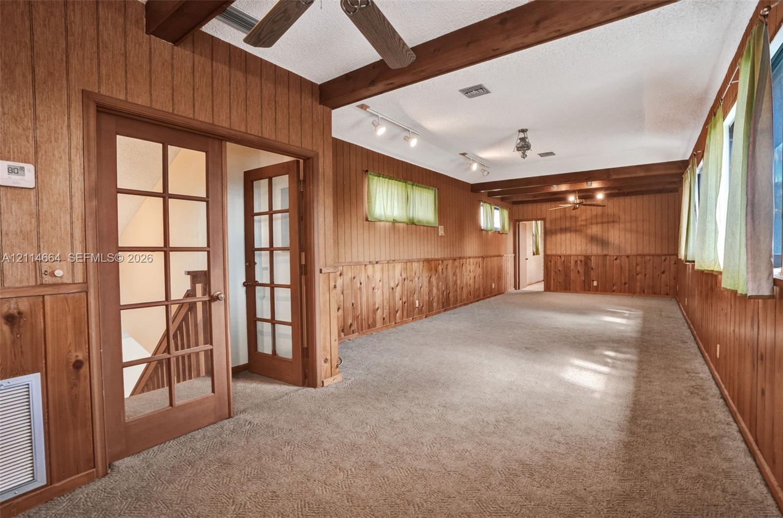 Wood-paneled interior featuring exposed structural ceiling beams, French doors with glass panes, wall-mounted track lighting, and ceiling fans