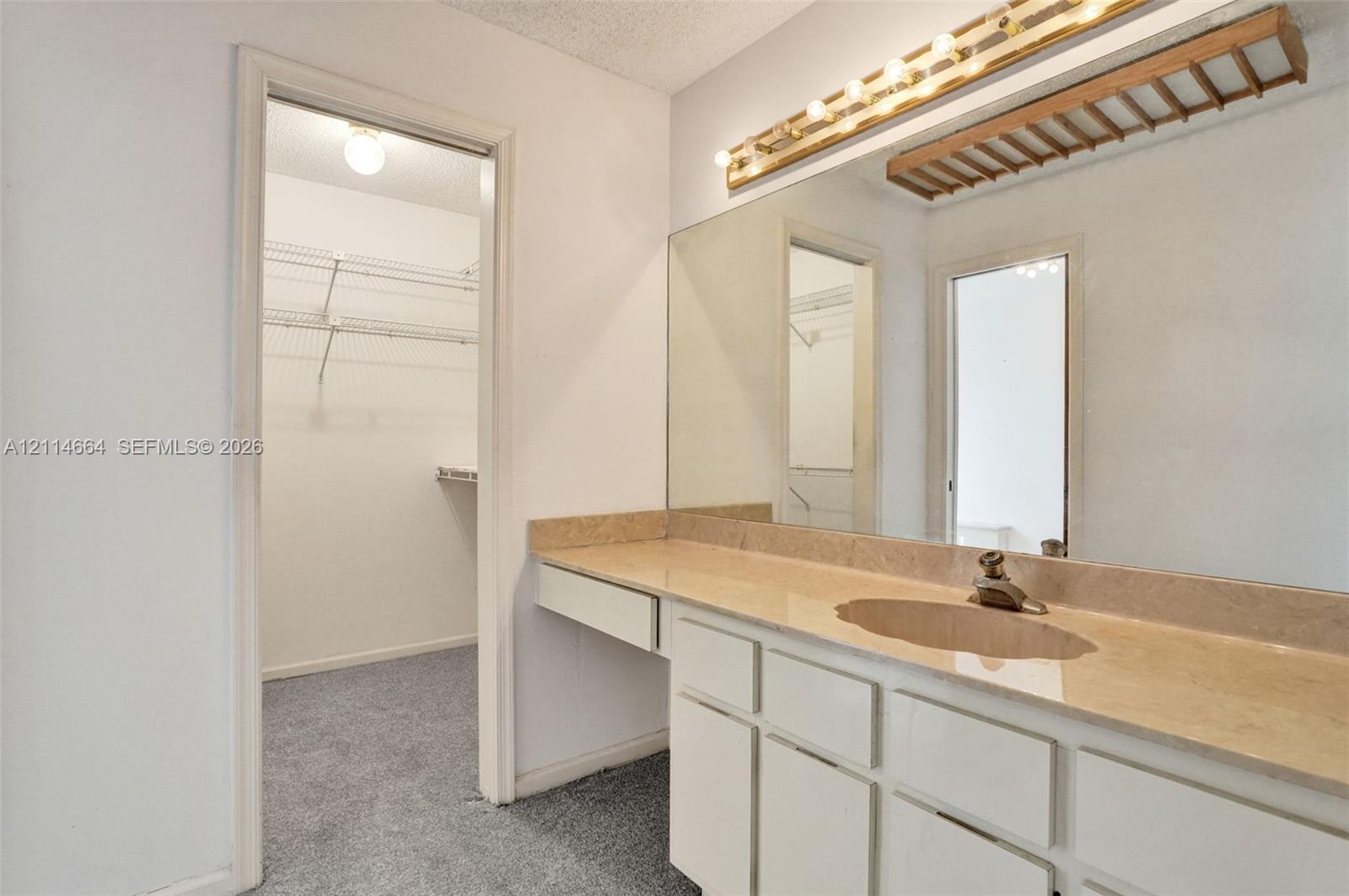 Vanity with neutral-toned solid surface countertop, integrated sink, and full-width mirror with mounted lighting, plus walk-in storage area featuring wire shelving