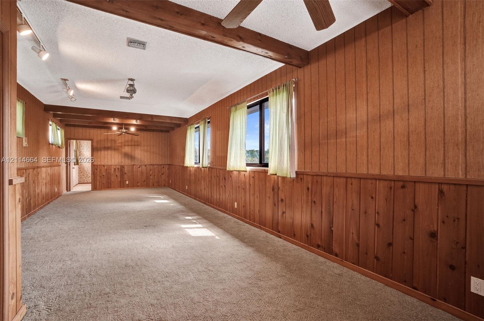 Wood-paneled walls, exposed timber ceiling beams, carpeted flooring, track lighting, and ceiling-mounted fans