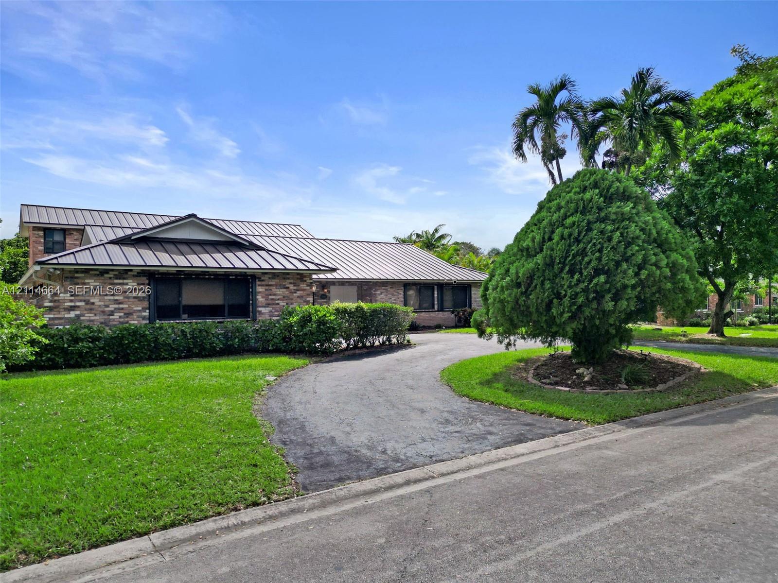 Single-story exterior featuring a brick facade, metal roofing, a curved paved driveway, and established landscaping