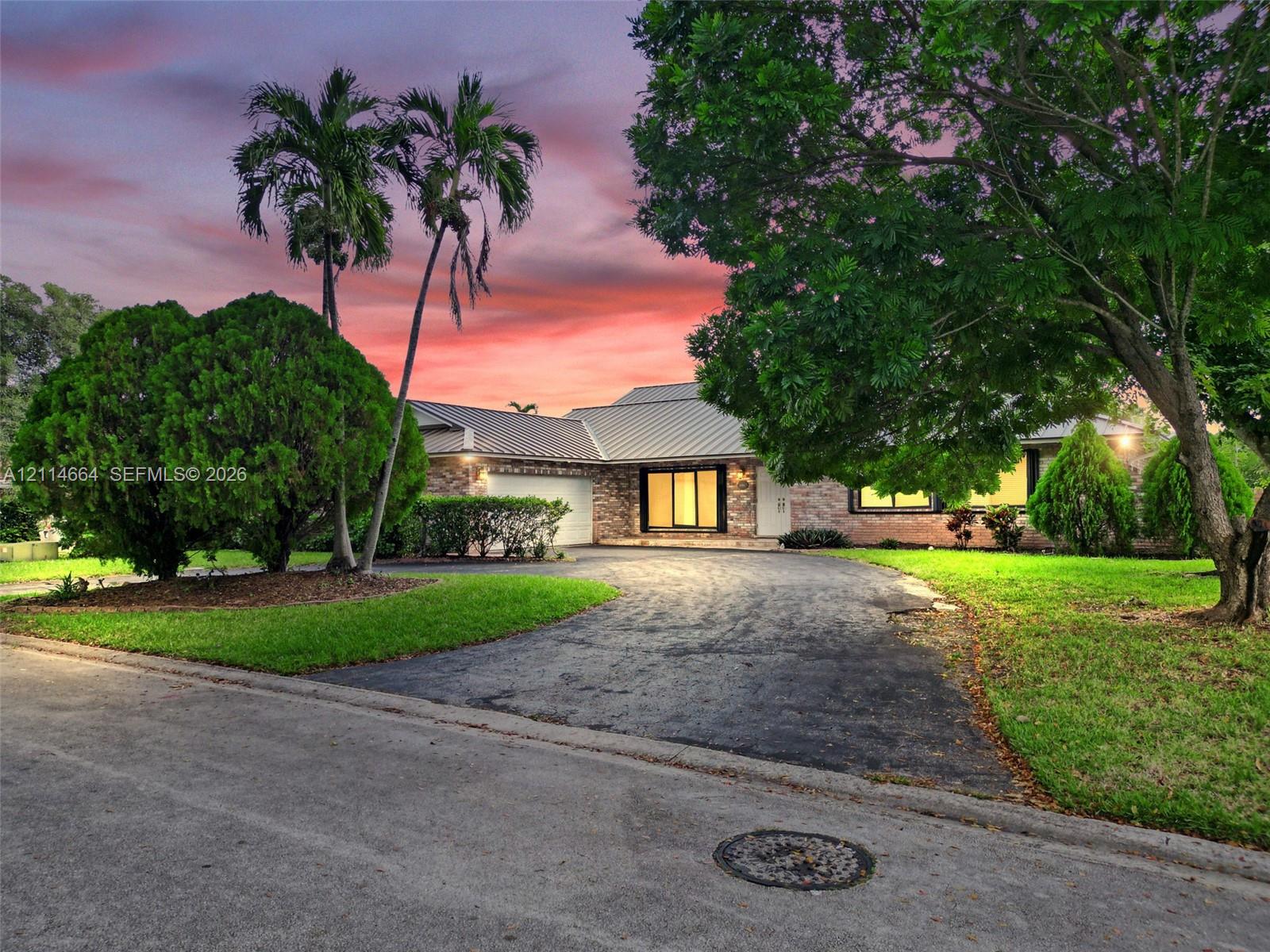 Single-story residence featuring a brick facade, metal roofing, paved driveway, and mature landscaping
