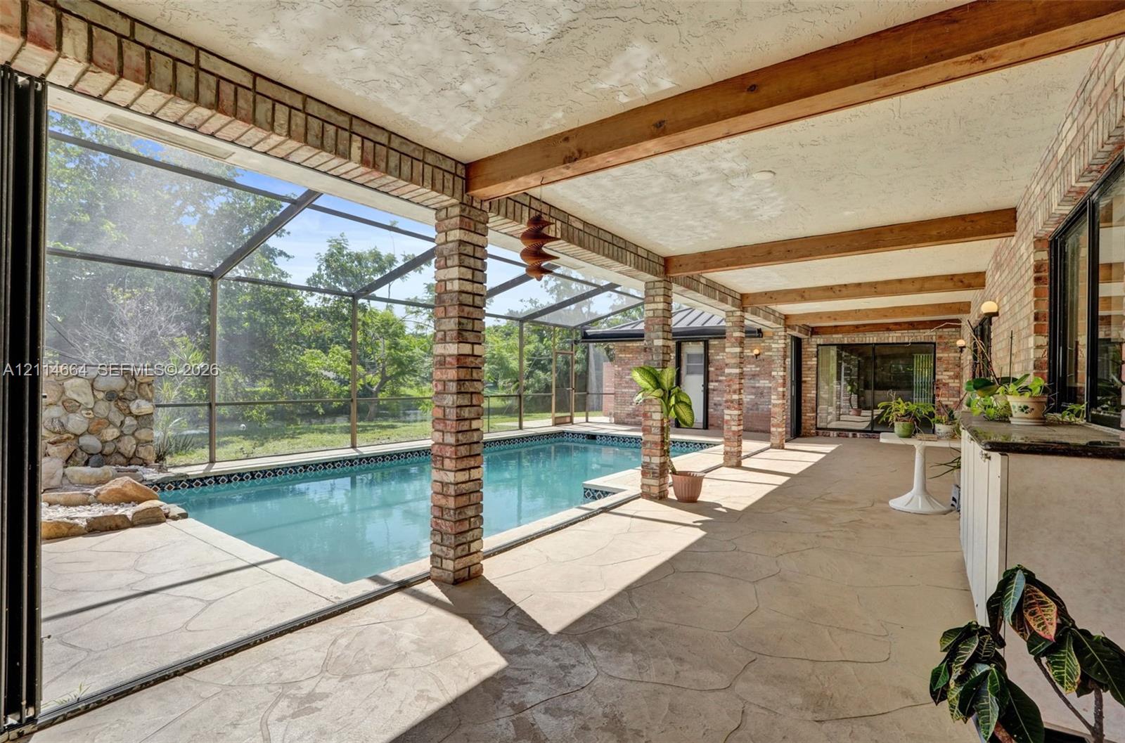 Covered patio featuring exposed wooden beams, brick support columns, a screened-in enclosure, and a swimming pool with a stone rock feature