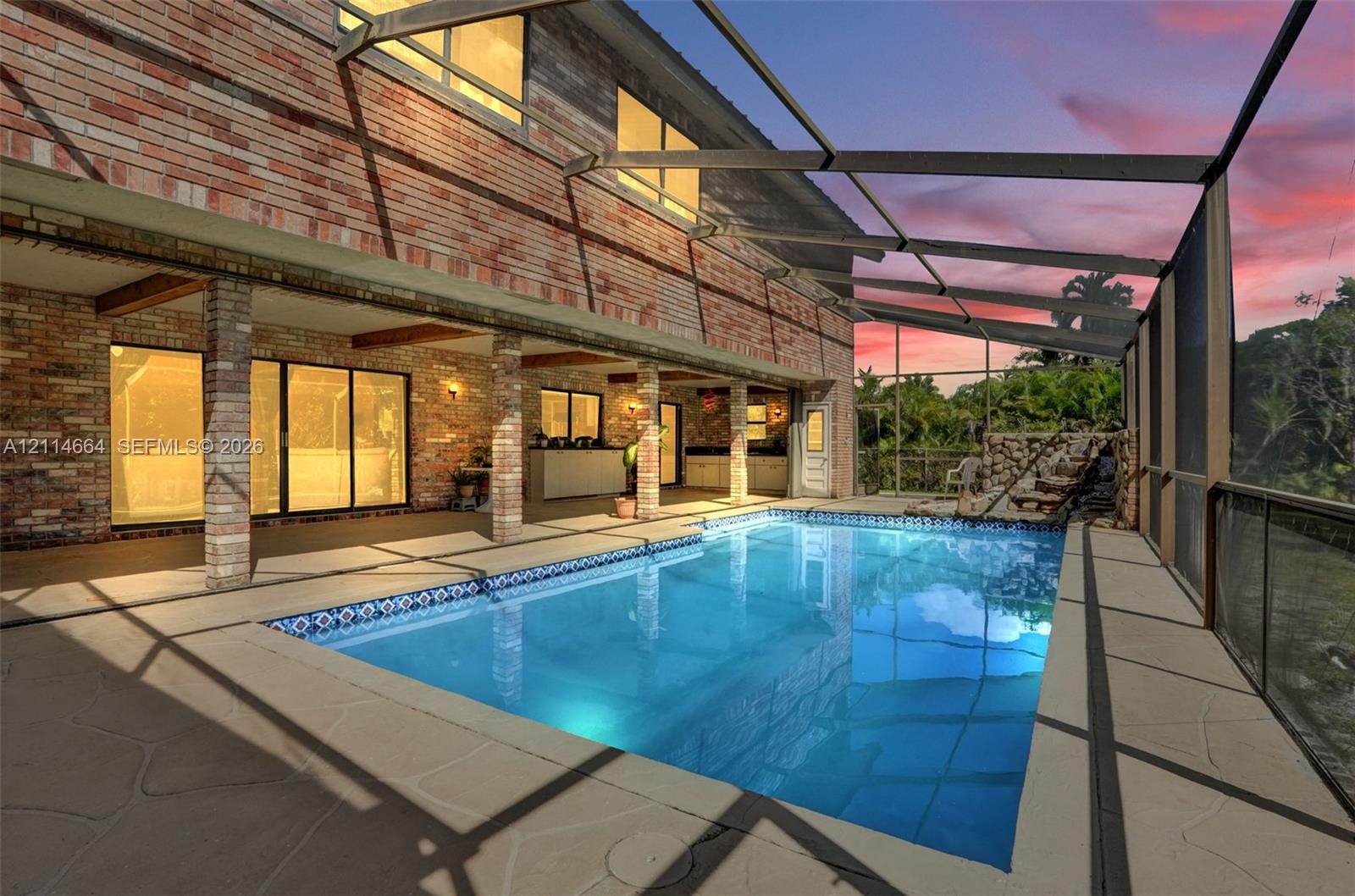 Screened pool enclosure featuring a brick facade, covered patio, rock-style water feature, and exterior masonry pillars