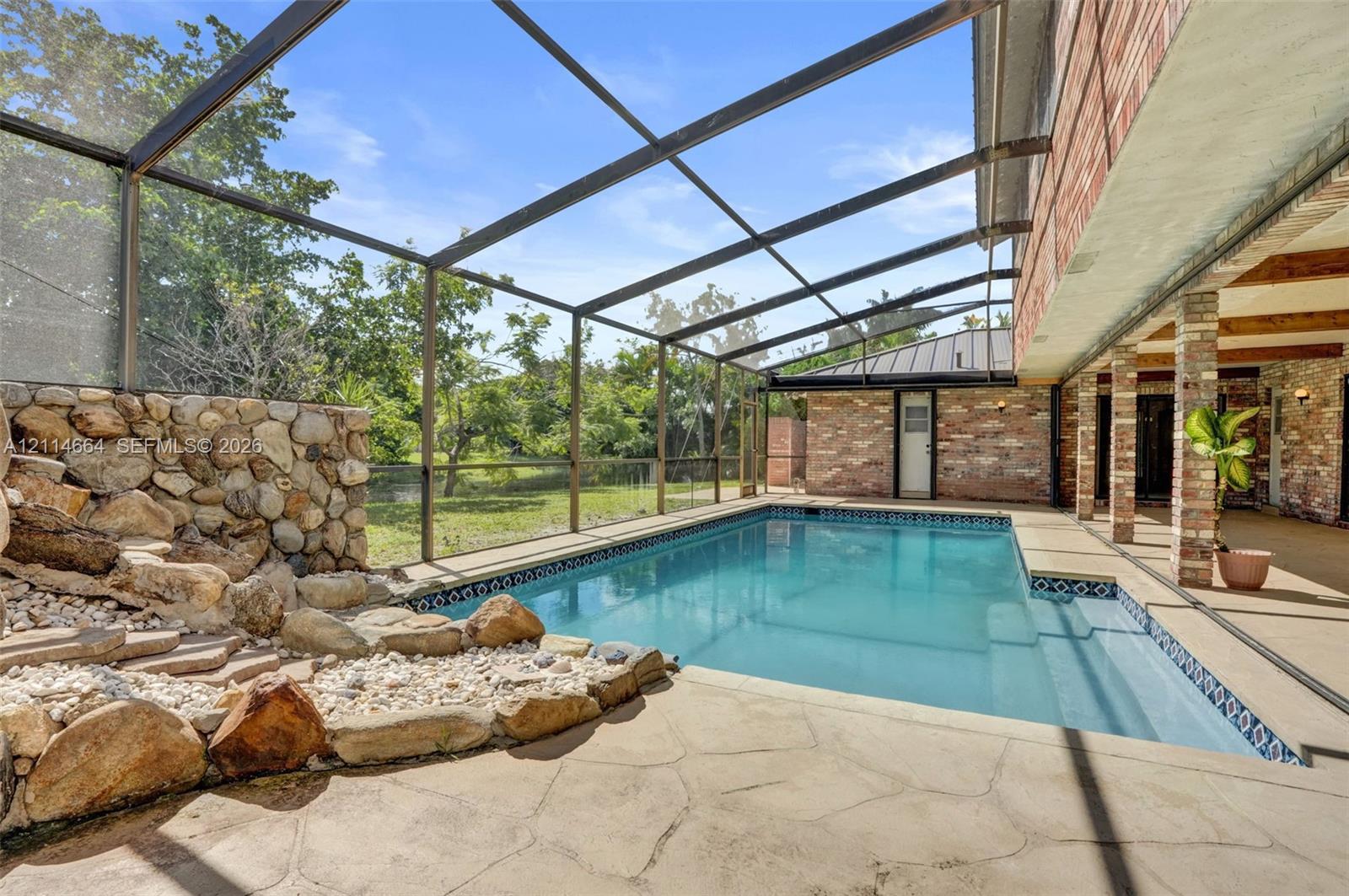 Screen-enclosed pool area featuring a natural stone water feature, patterned concrete decking, covered patio space, and brick architectural detailing