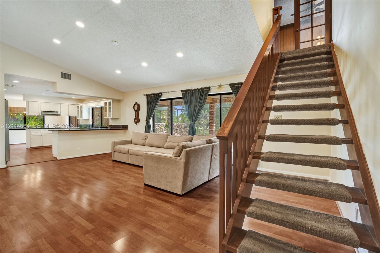 Open-concept living space featuring wood-finish flooring, vaulted ceilings with recessed lighting, a wooden staircase with carpeted treads, and sliding glass doors