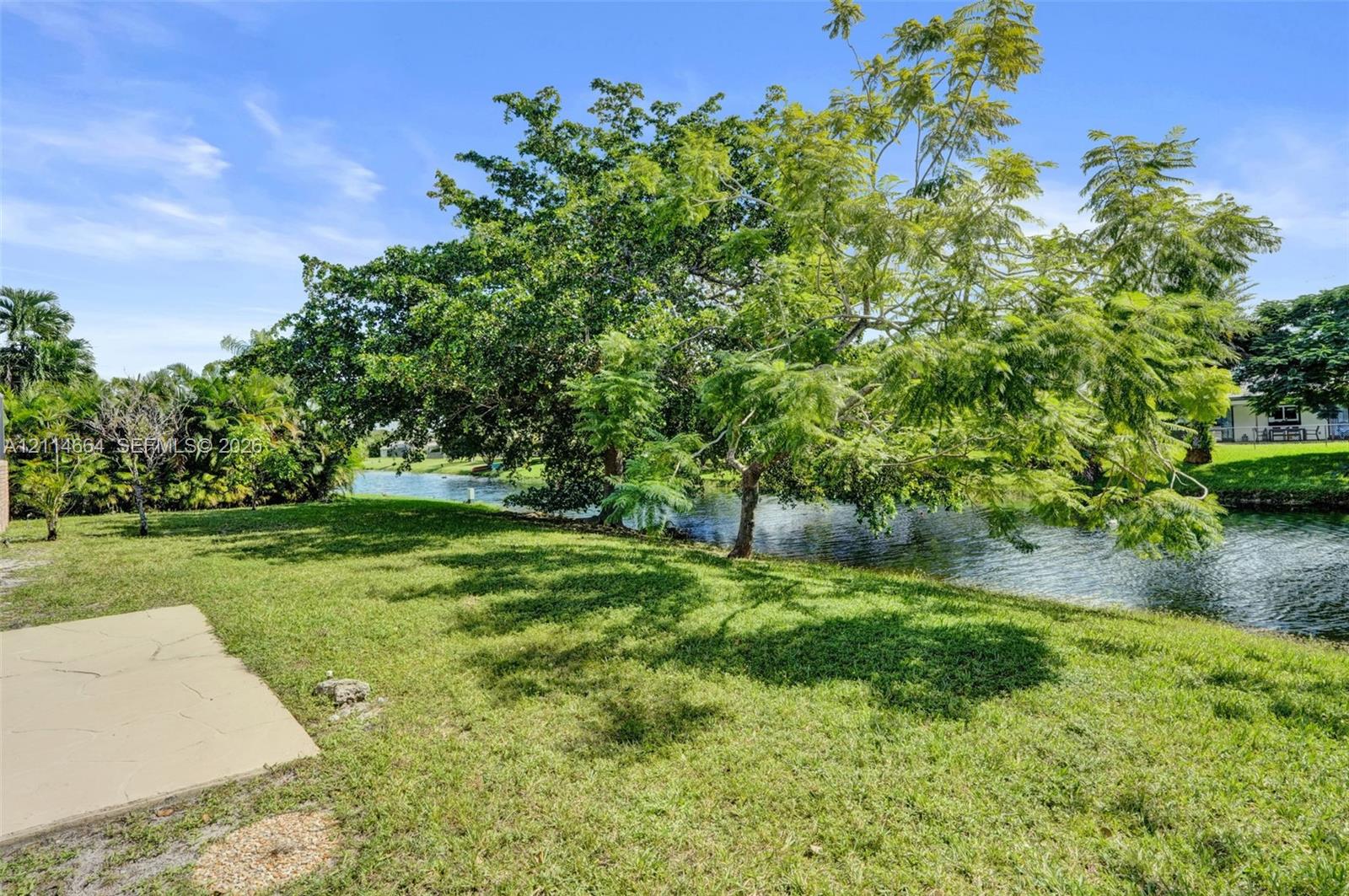 Rear grounds featuring a waterfront lot, concrete hardscaping, manicured lawn, and mature trees