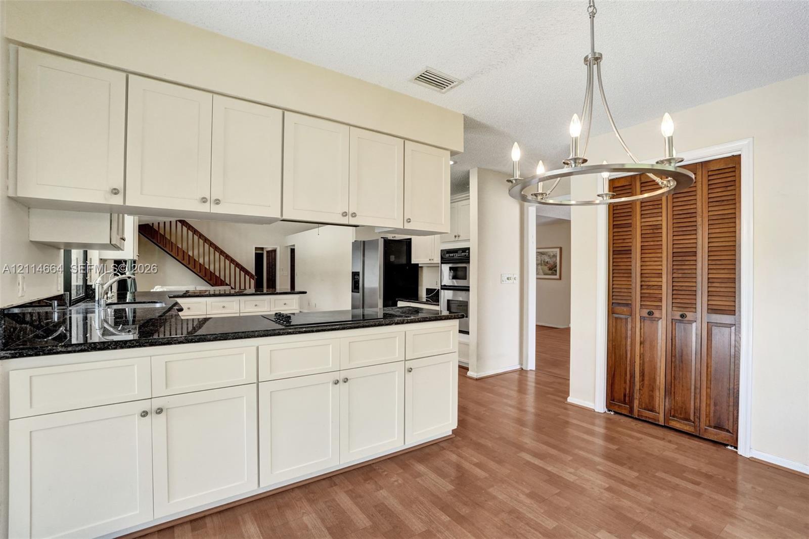 Kitchen featuring white shaker-style cabinetry, dark stone-finish countertops, stainless steel wall-mounted oven, wood-finish flooring, and a circular metal chandelier