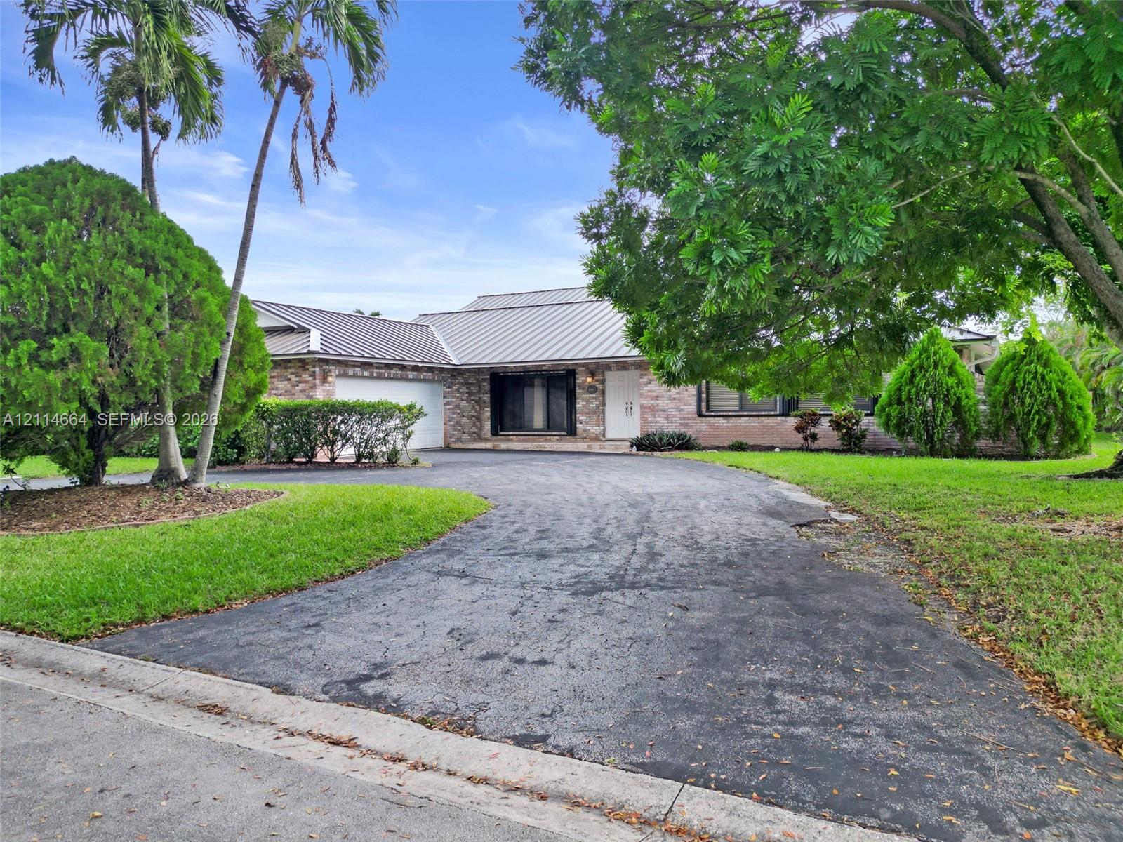 Single-story brick exterior featuring a multi-pitched metal roof, attached garage, and paved driveway surrounded by mature landscaping and established trees
