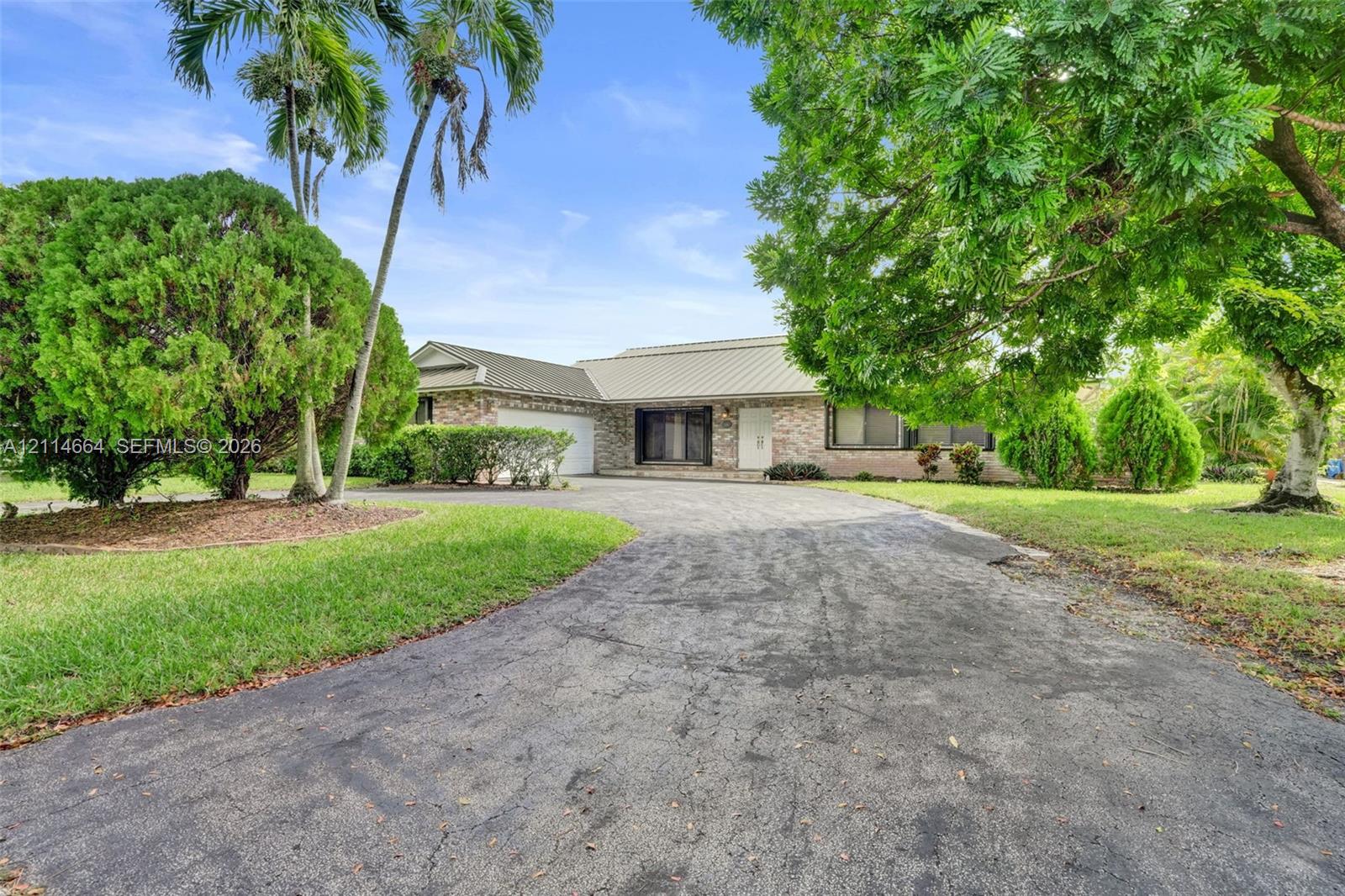 Single-story residence featuring a brick facade, multi-gabled metal roof, attached garage, and mature palm trees situated on a landscaped lot