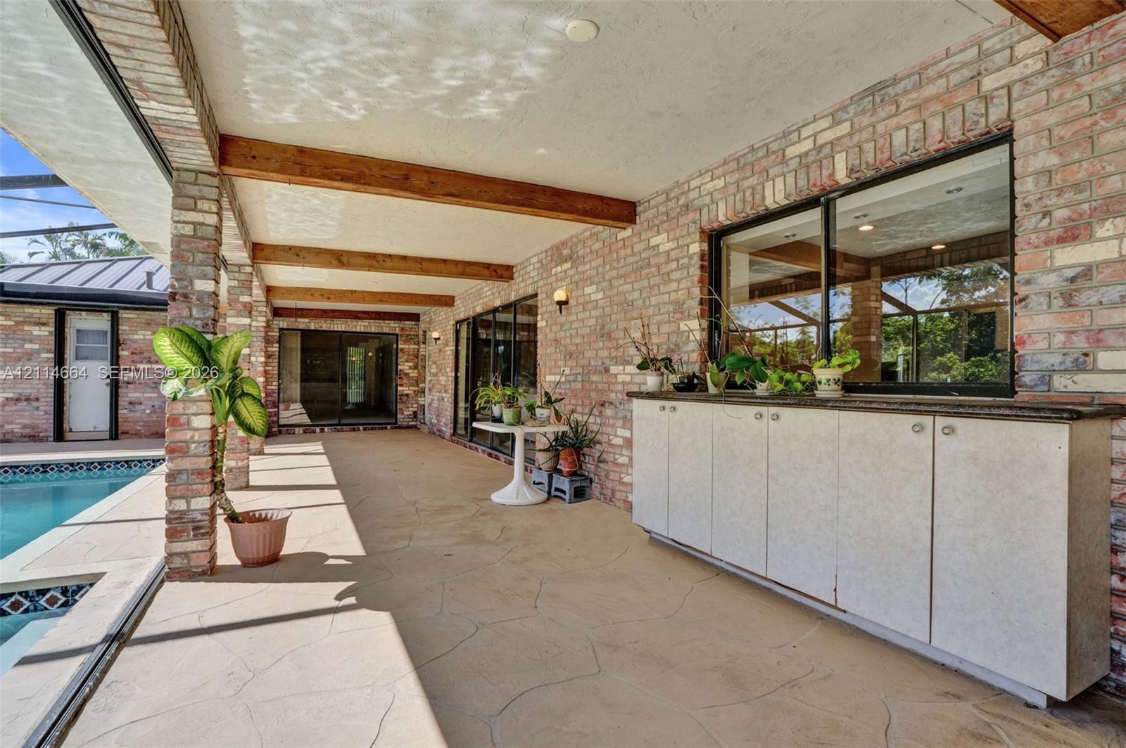 Covered outdoor patio featuring exposed wood beams, brick masonry walls, stamped concrete flooring, and built-in cabinetry with a stone countertop