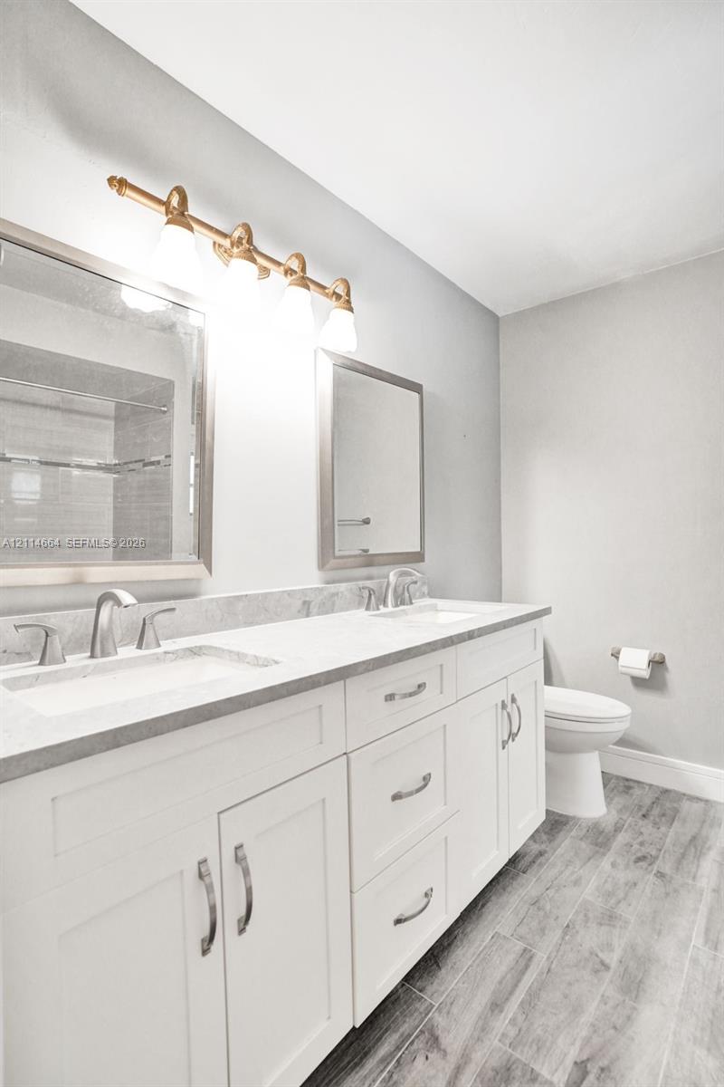 Dual-sink vanity with stone countertop, white shaker-style cabinetry, metallic hardware, and wood-finish flooring