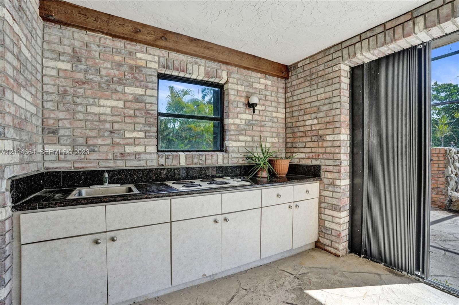 Outdoor kitchen area featuring exposed brick walls, stone flooring, built-in cabinetry, dark stone-finish countertops, and an integrated cooktop and sink