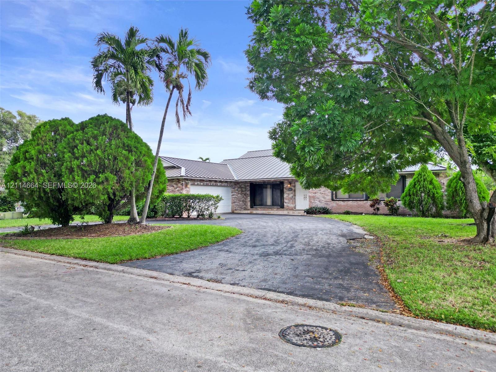 Single-story brick exterior featuring a metal roof, attached two-car garage, mature landscaping, and an expansive paved driveway