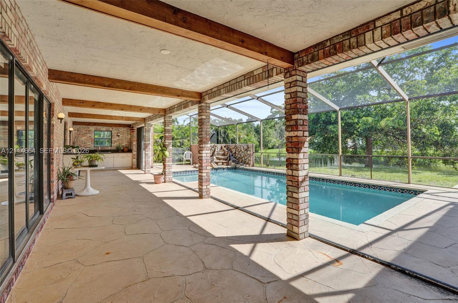 Covered lanai with flagstone flooring, exposed wood ceiling beams, brick support columns, and an in-ground pool enclosed by a screen structure