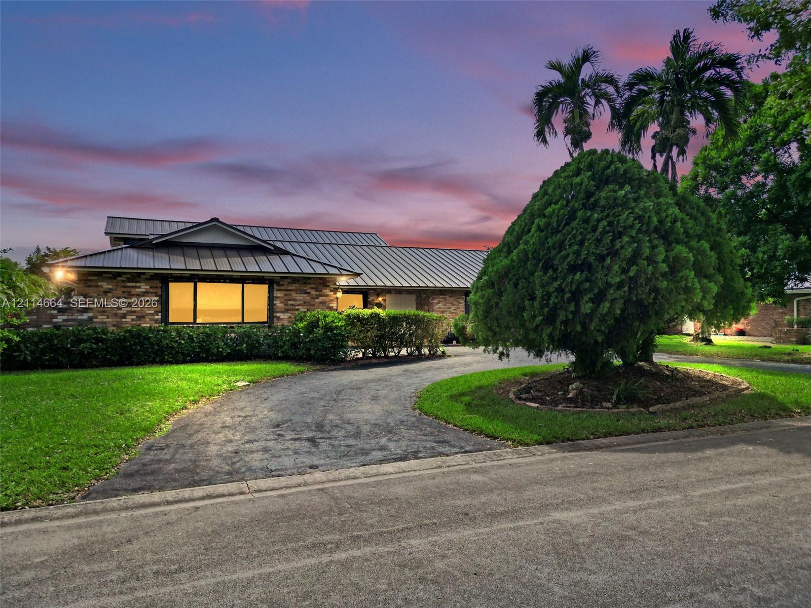 Single-story residence featuring a multi-pitched metal roof, stone veneer exterior, expansive front lawn, circular asphalt driveway, and mature landscaping