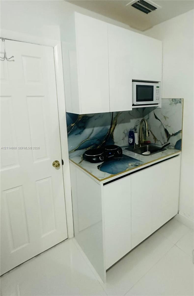 Compact kitchenette featuring white flat-panel cabinetry, patterned stone-style backsplash, integrated microwave, drop-in sink with a high-arch faucet, and polished flooring