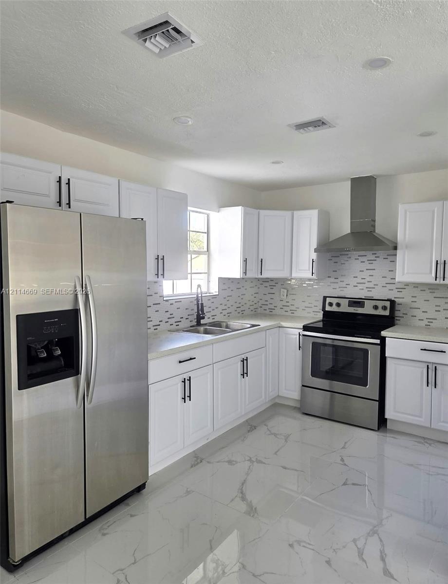 Kitchen featuring white shaker cabinetry, stainless steel appliances, a range hood, decorative tile backsplash, and polished stone-finish flooring Kitchen featuring white shaker cabinetry, stainless steel appliances, a range hood, decorative tile backsplash, and polished stone-finish flooring