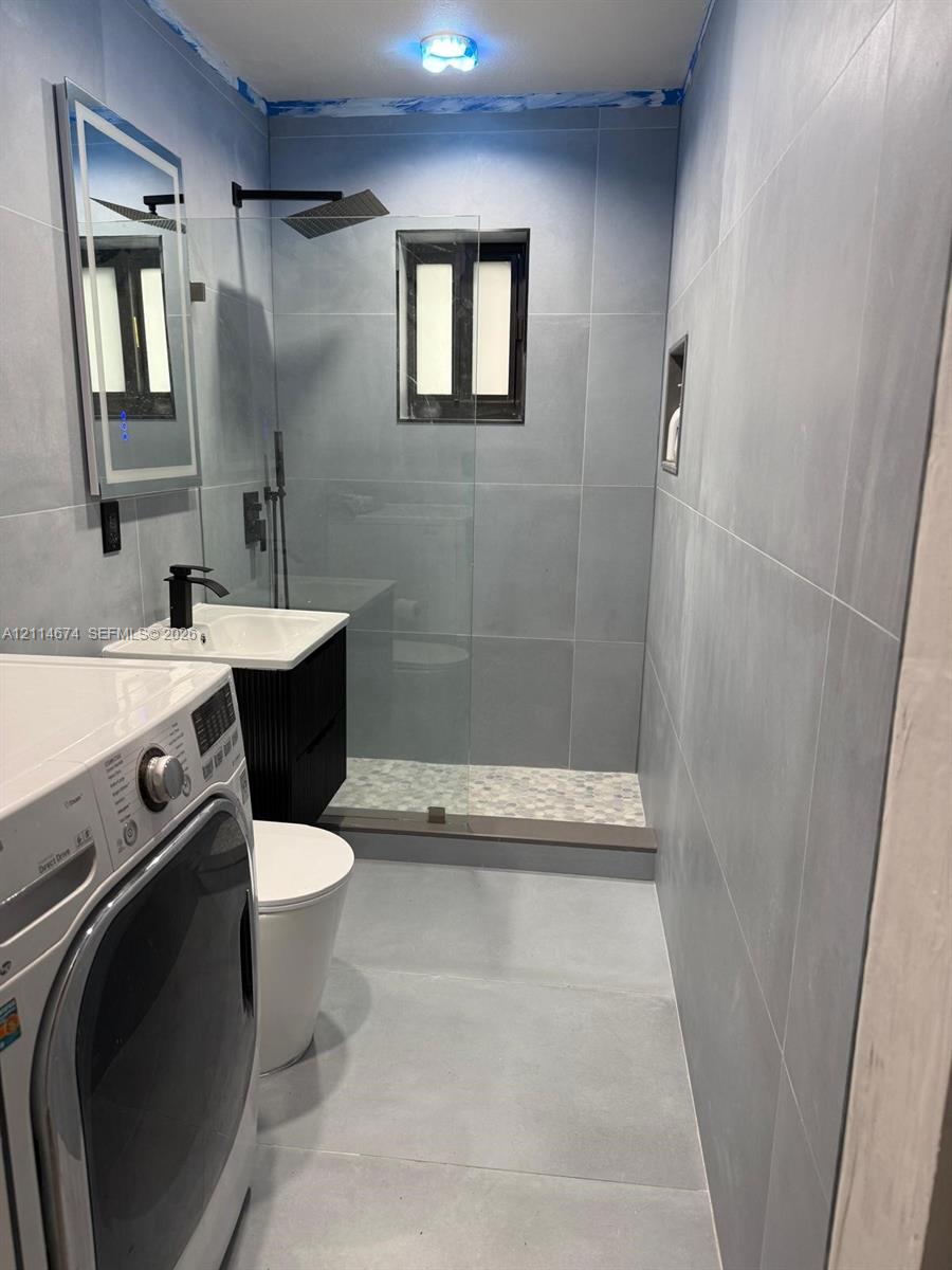 Walk-in shower with a glass panel, stone-finish shower floor, matte black rainfall showerhead, wall-mounted vanity with a rectangular sink, and illuminated wall mirror Walk-in shower with a glass panel, stone-finish shower floor, matte black rainfall showerhead, wall-mounted vanity with a rectangular sink, and illuminated wall mirror