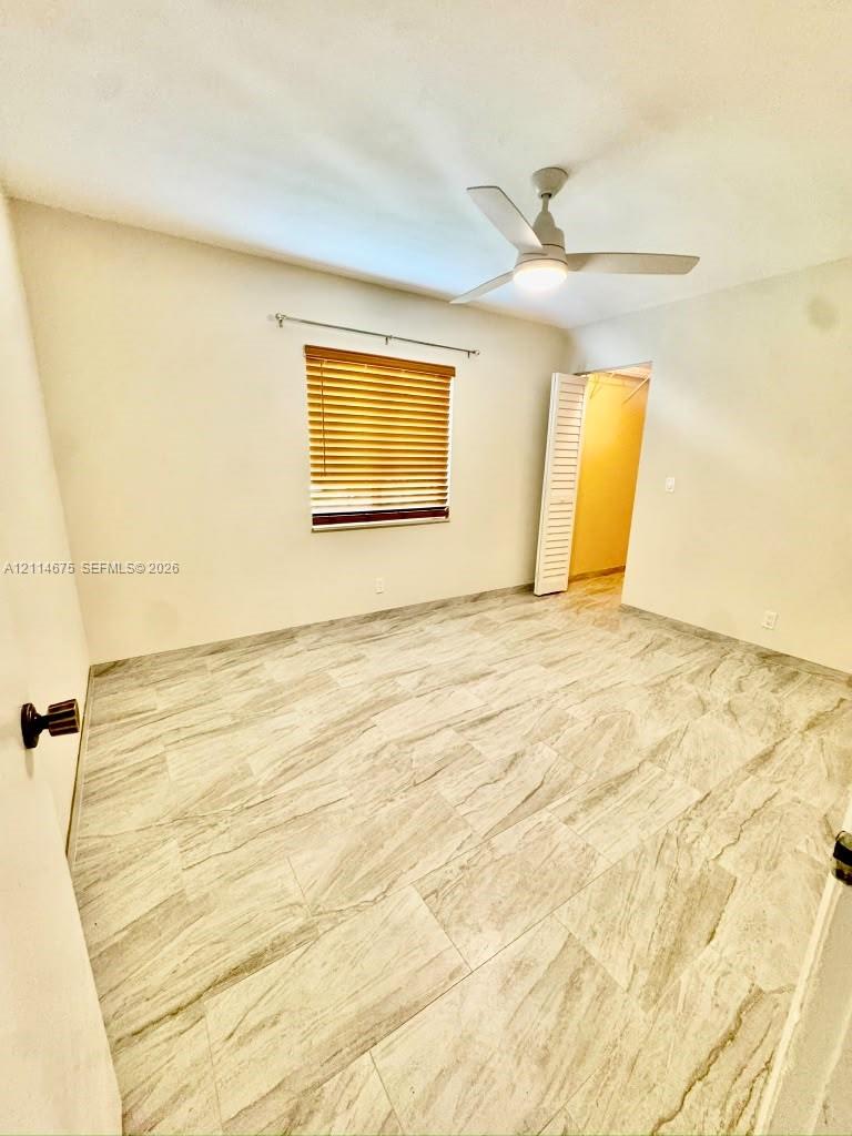 Neutral-toned interior featuring large-format stone-patterned tile flooring, a centered ceiling fan with integrated light fixture, window with horizontal blinds, and a louvered bi-fold closet door