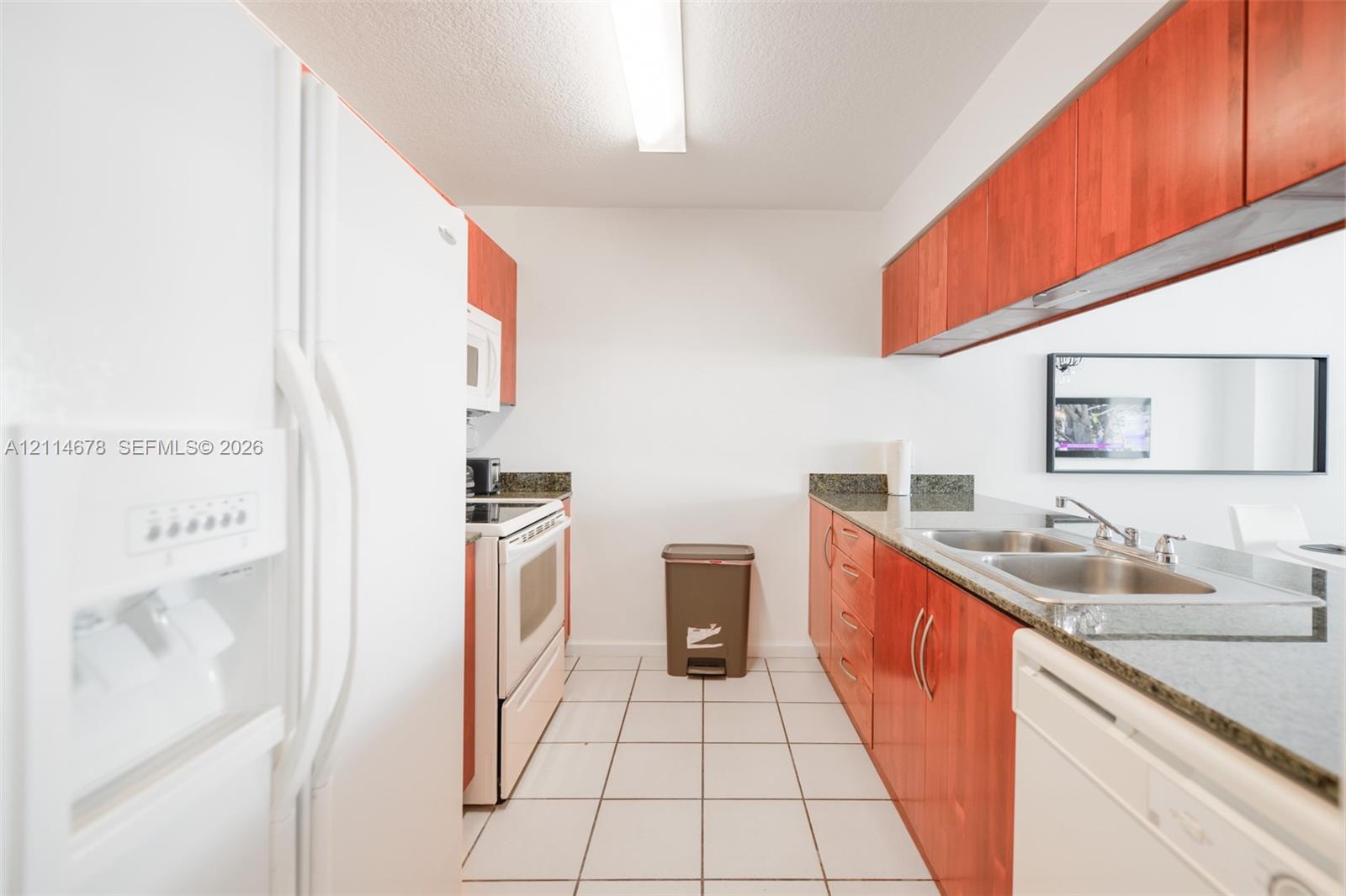 Kitchen featuring wood-finish cabinetry, stone-surface countertops, dual-basin stainless steel sink, tile flooring, and integrated white appliances