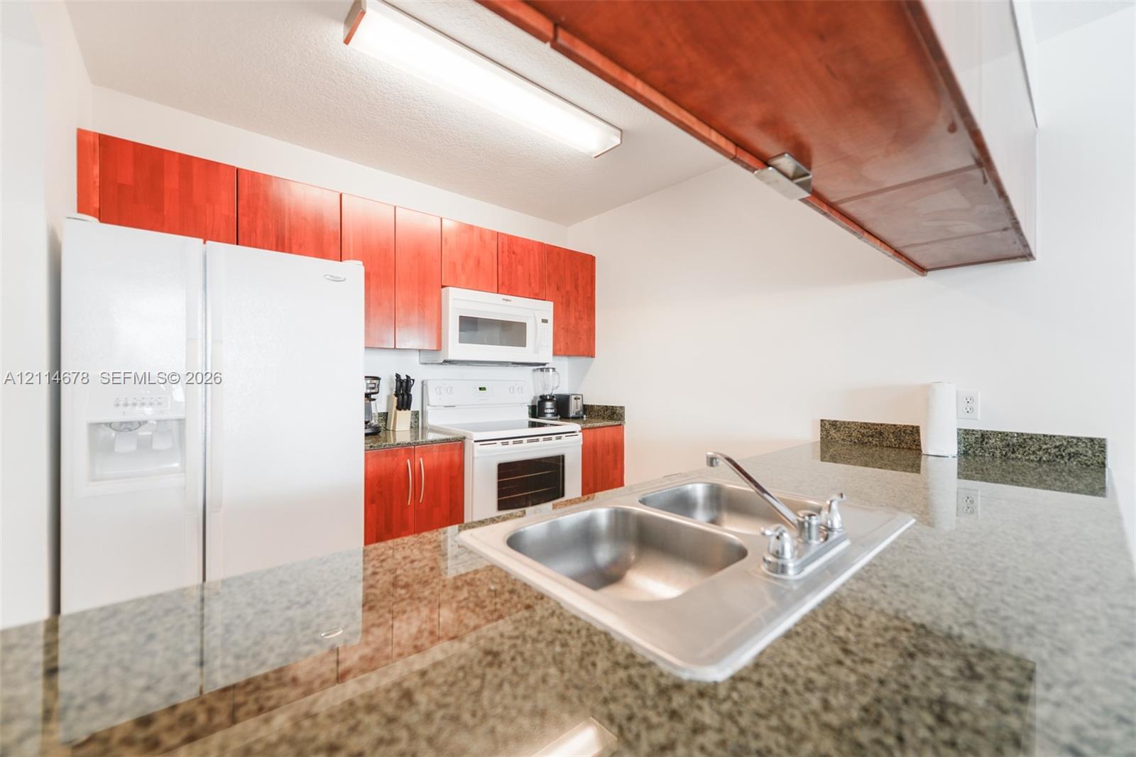 Kitchen featuring stone-finish countertops, stainless steel double-basin sink with a chrome faucet, wood-finish cabinetry, and built-in appliances including a refrigerator, microwave, and range