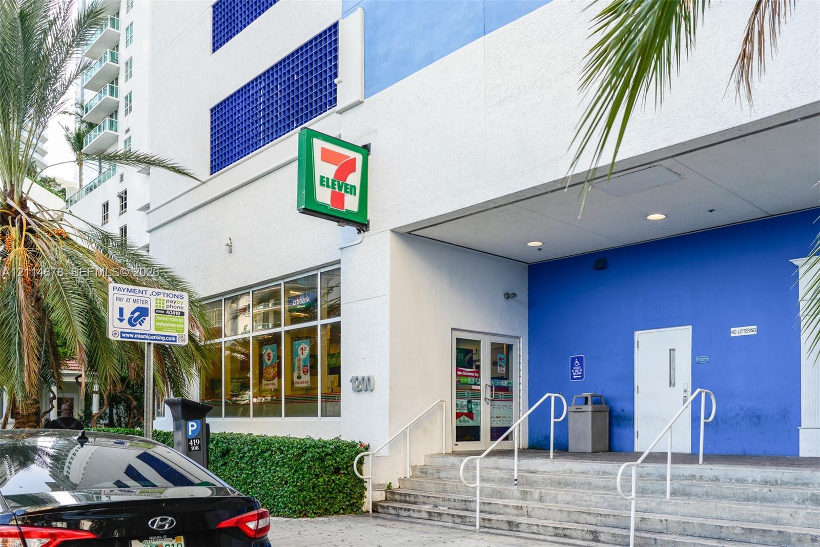 Multi-story exterior featuring white and blue facade, recessed entryway with concrete stairs, metal handrails, and glass-paneled storefront doors