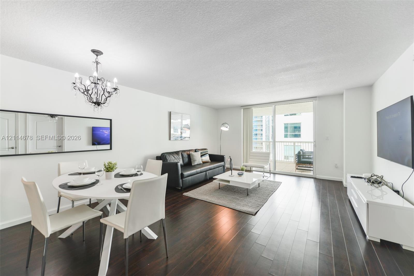 Open-concept living space featuring wood-finish flooring, a contemporary multi-light chandelier, a large wall-mounted mirror, and sliding glass doors providing access to a private balcony