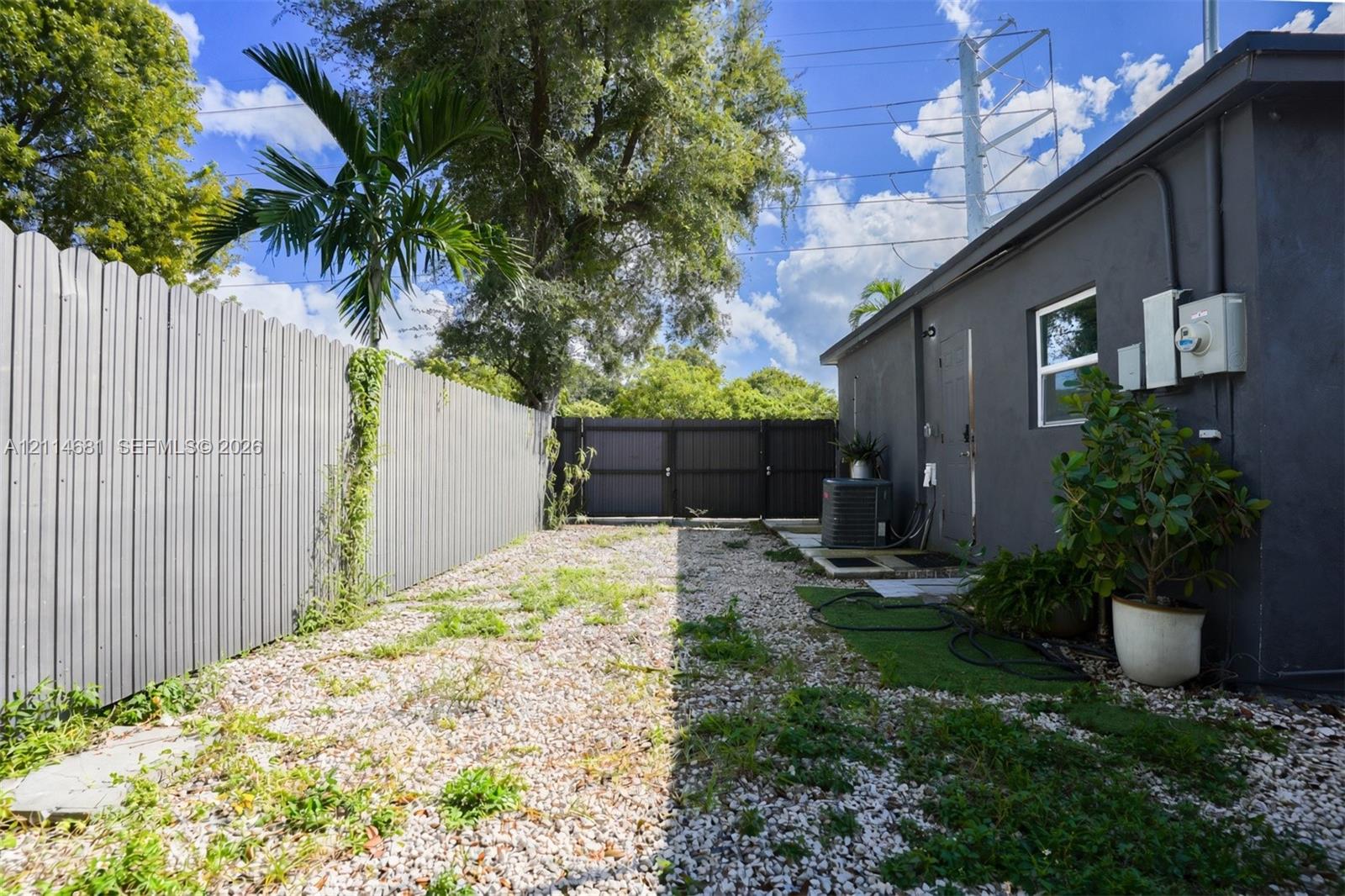 Side yard enclosed by corrugated metal fencing, featuring a gravel surface, palm tree, and gated entry