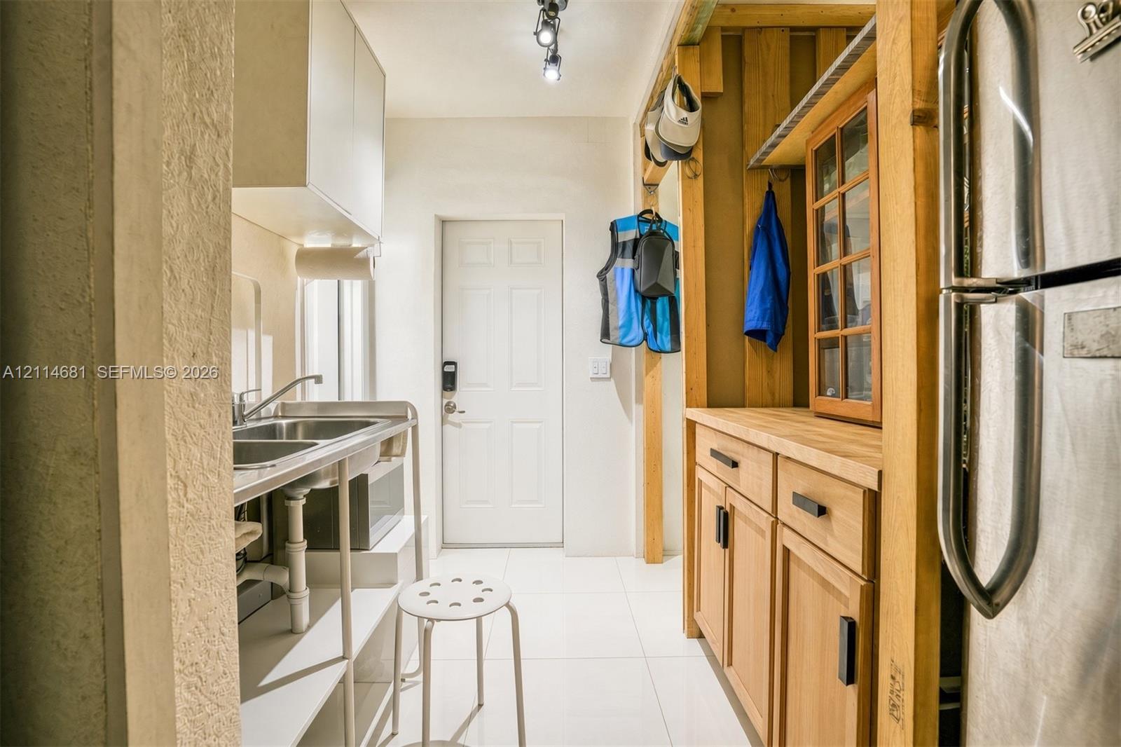 Stainless steel double-basin sink, light wood cabinetry with matte black hardware, butcher-block style countertop, white tile flooring, and track lighting
