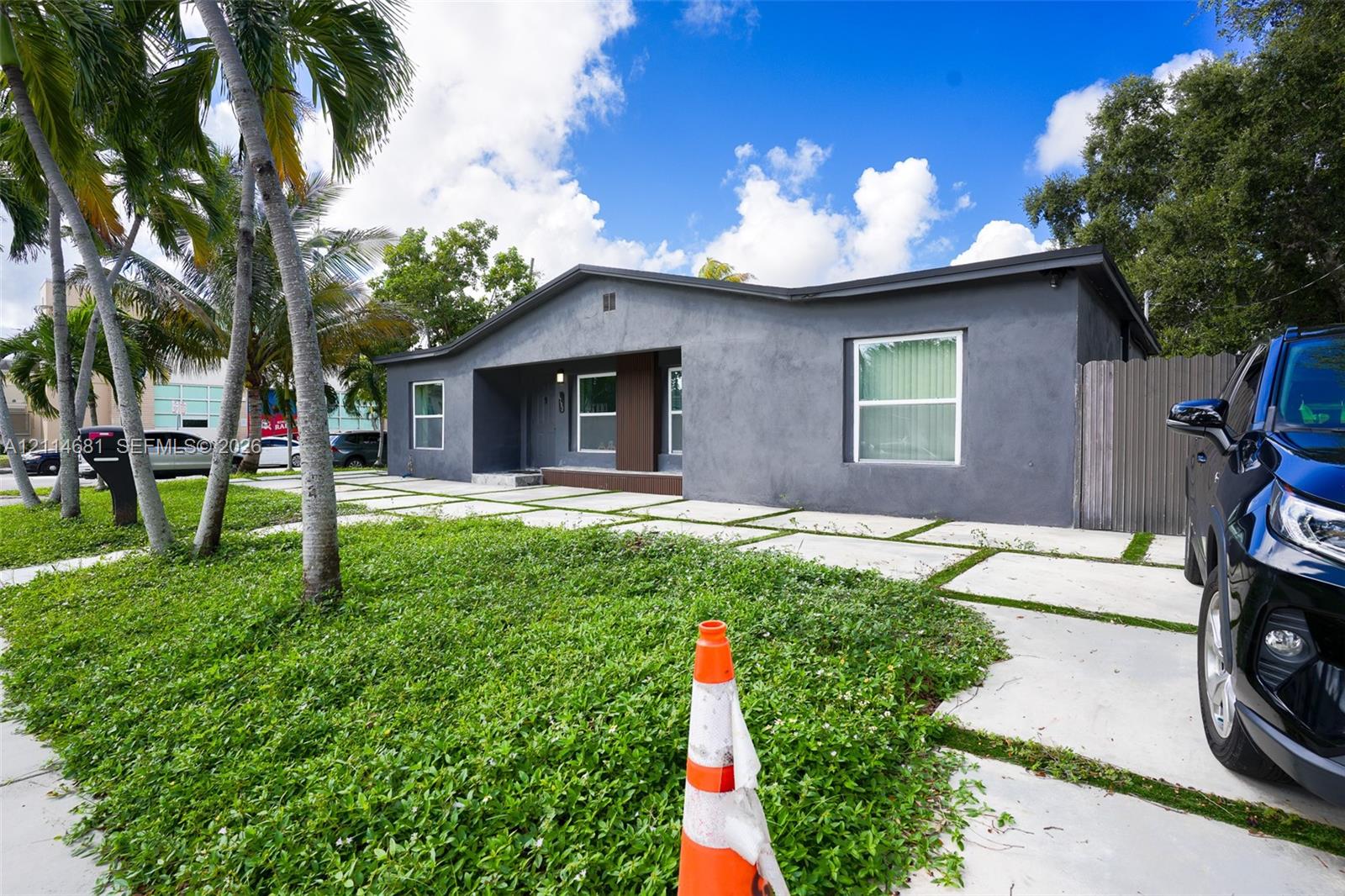 Single-story exterior featuring a dark-toned stucco facade, a grid-pattern concrete paver driveway, and a covered front entry with wood-texture paneling