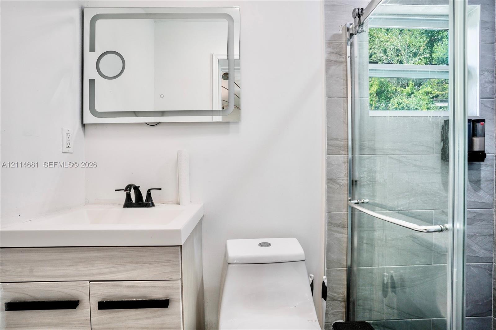 Vanity with a white integrated sink, wood-finish cabinetry, matte black hardware, backlit mirror, and a walk-in shower featuring grey tile surround and a sliding glass door