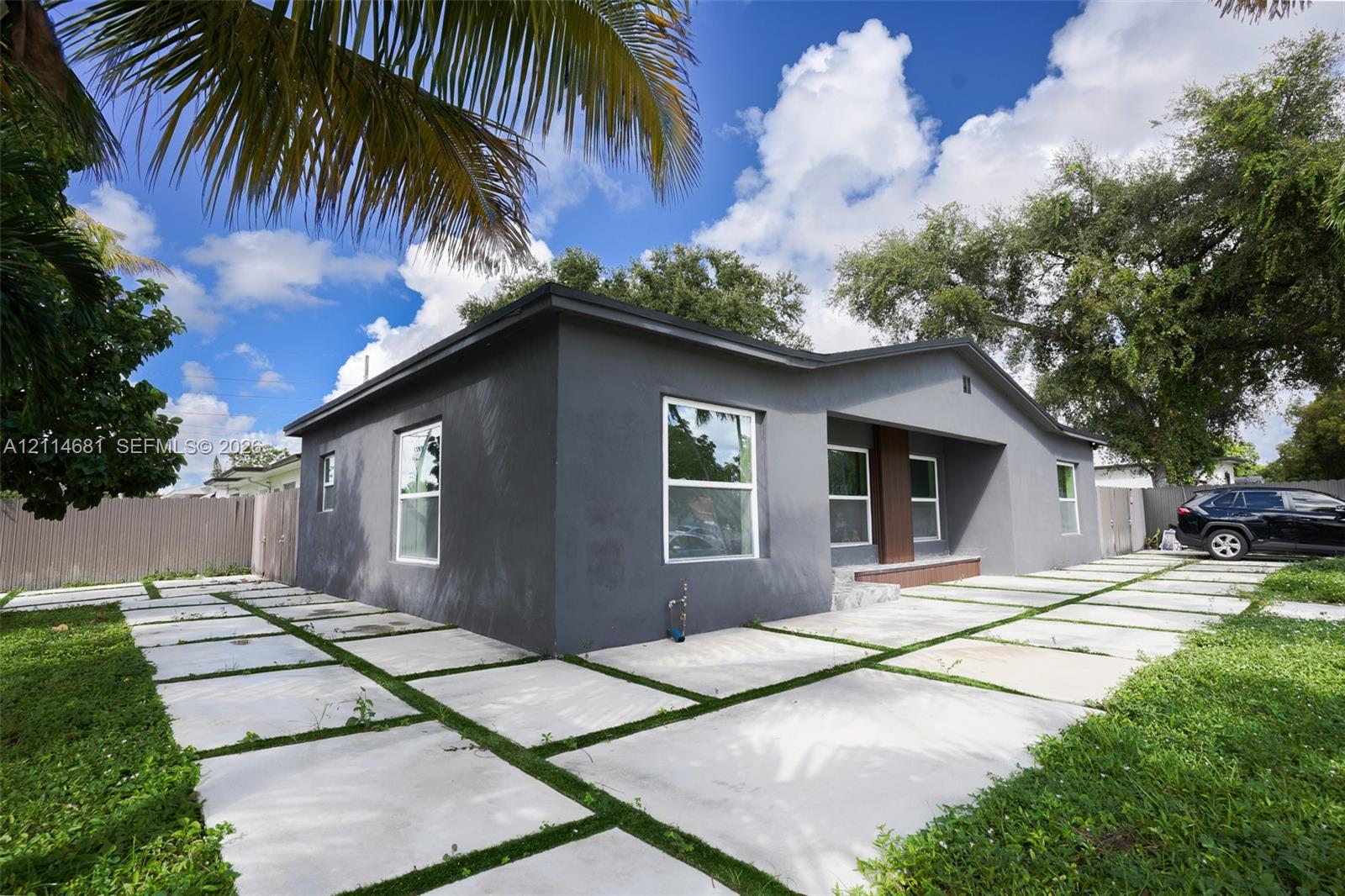 Single-story exterior featuring a dark-toned stucco facade, grid-patterned concrete paver driveway, and a recessed entryway with a wooden accent pillar
