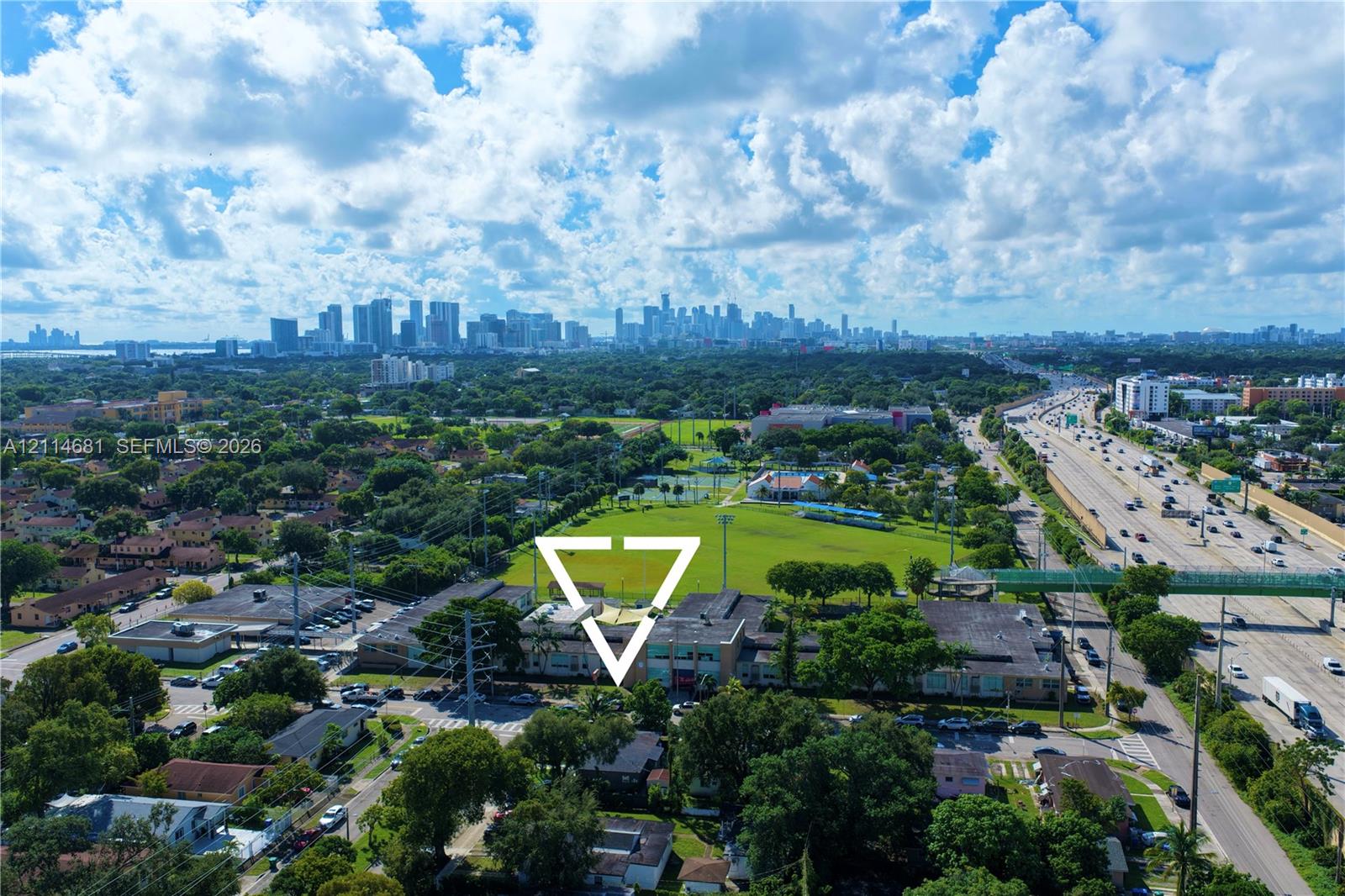 Aerial perspective of the property grounds featuring a multi-story building, established mature trees, proximity to expansive green space, and a backdrop of the urban skyline