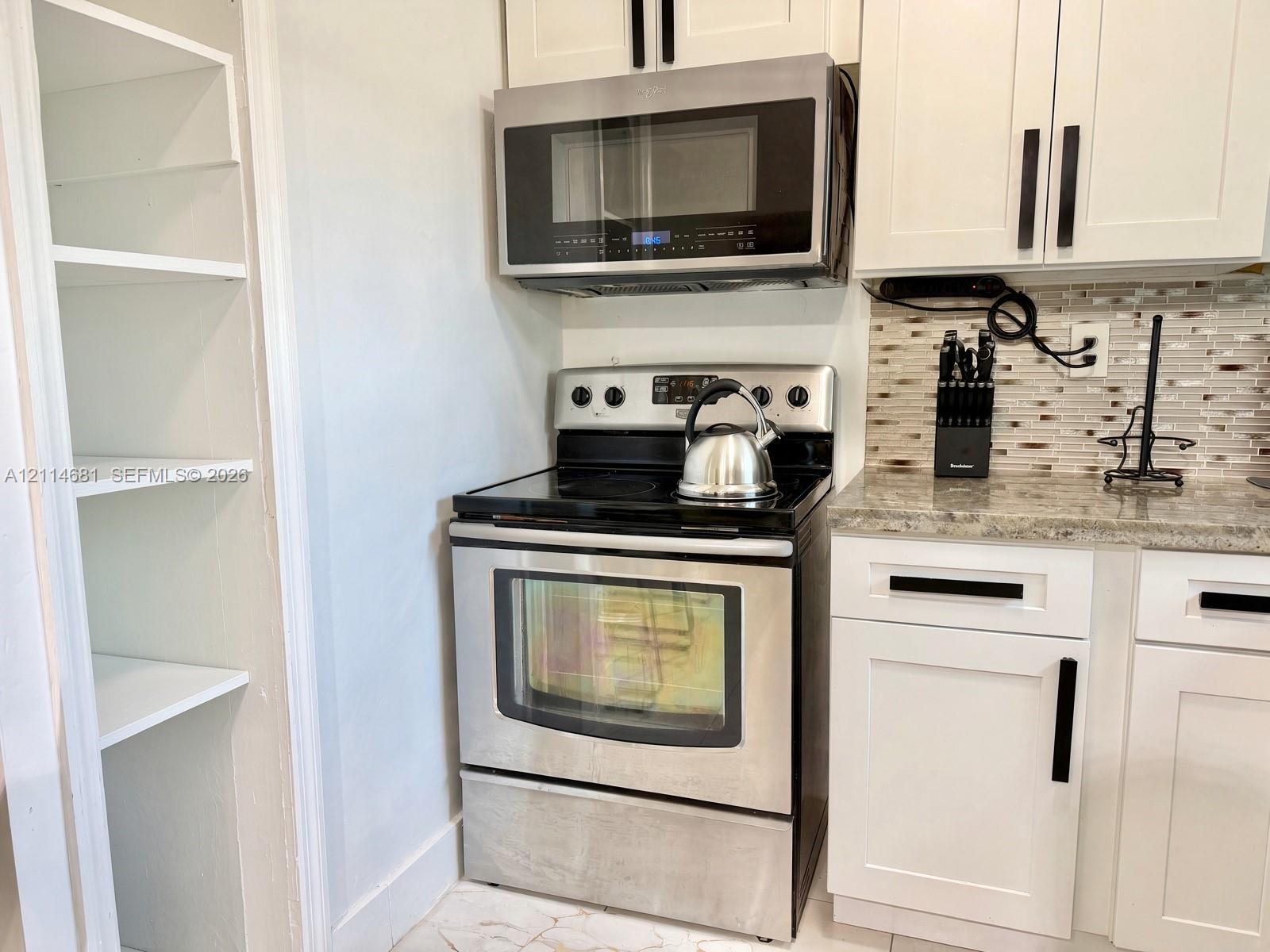 Kitchen featuring white shaker-style cabinetry with matte black hardware, stainless steel appliances, a multi-tonal tile backsplash, stone-finish countertops, and an integrated open shelving pantry