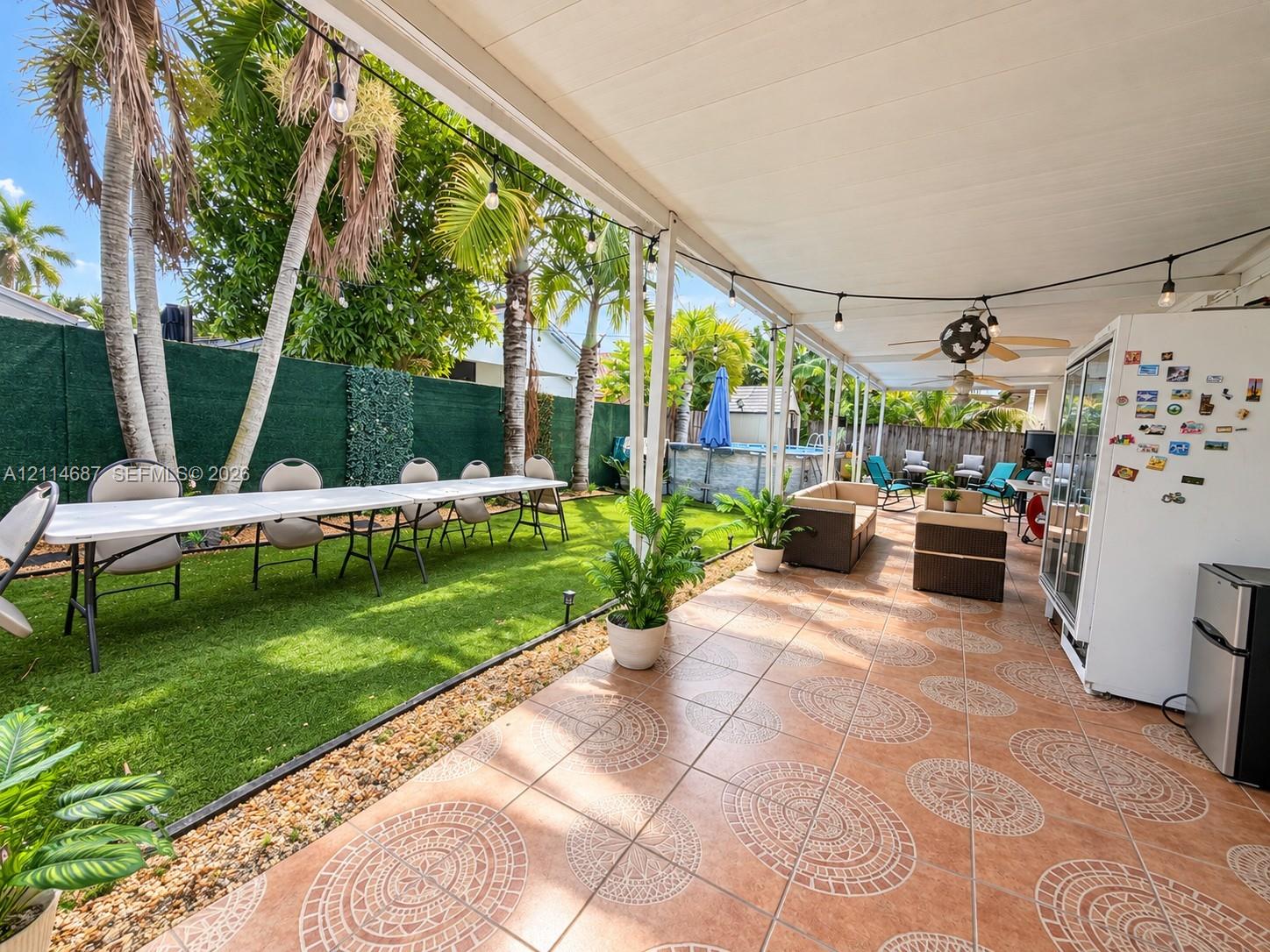 Covered patio featuring patterned tile flooring, ceiling fans, and string lighting, complemented by an artificial turf lawn and perimeter fencing
