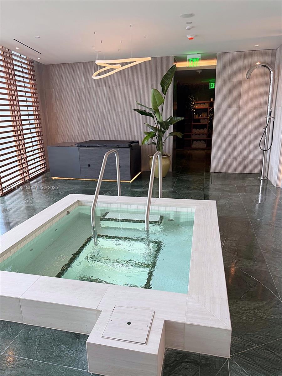 Indoor plunge pool featuring a stone-tiled surround, polished metal handrails, dark stone-finish flooring, linear pendant lighting, and a wall-mounted shower fixture